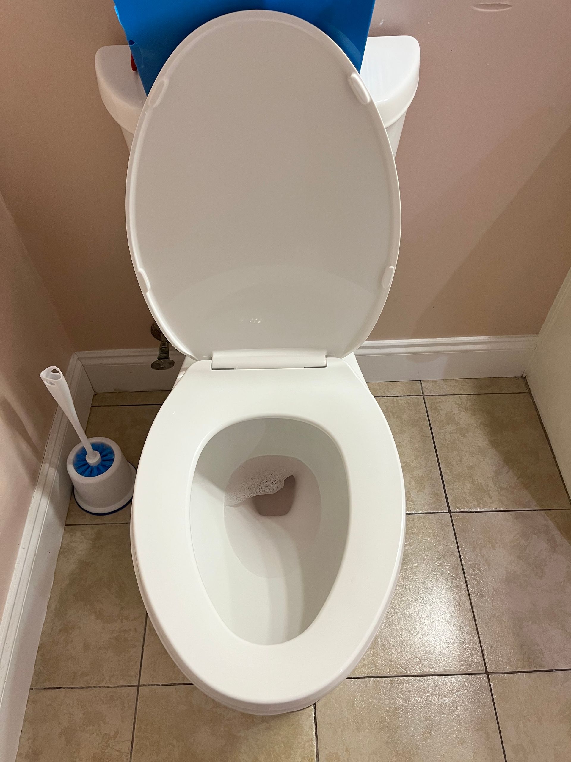 A white toilet with the seat up in a bathroom with tan tile floors and a toilet brush next to it.
