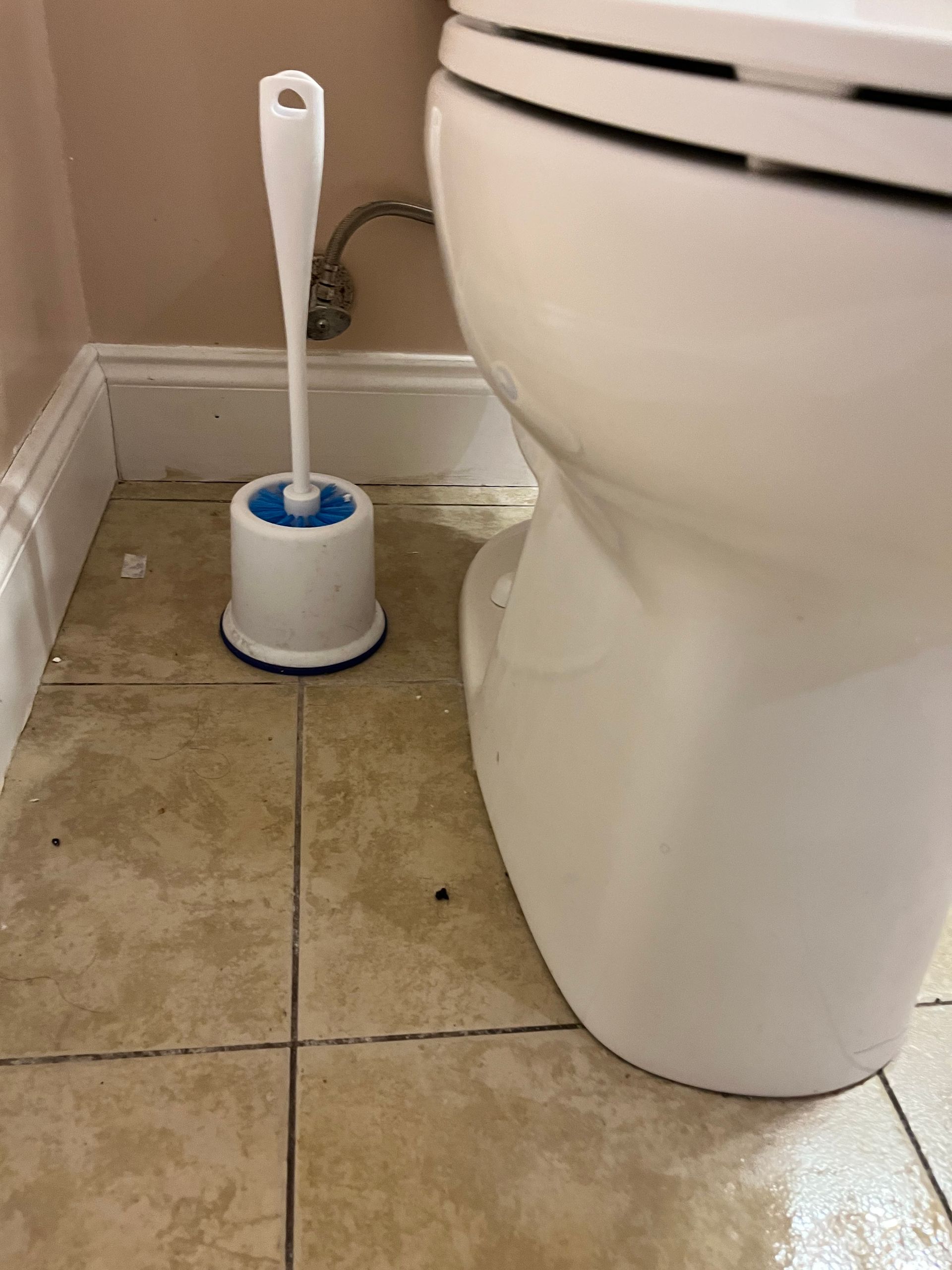 White toilet brush and holder next to a white toilet on tan tiled floor.