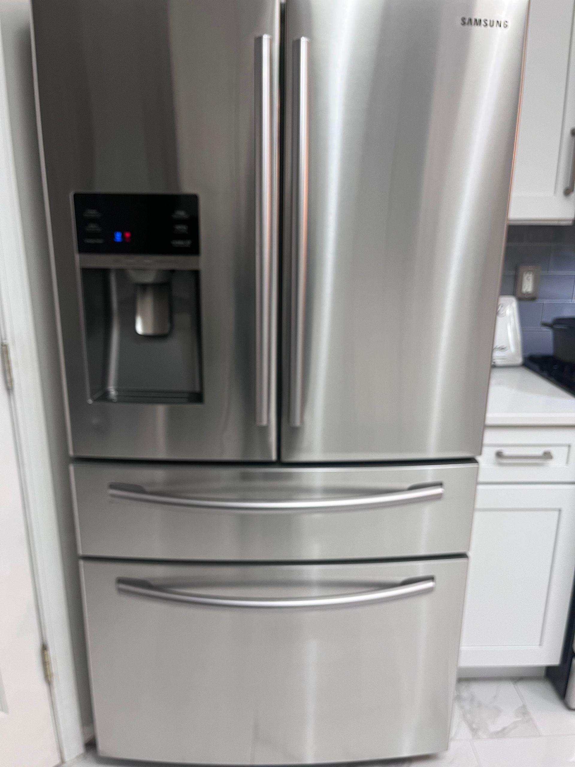 Stainless steel Samsung refrigerator with a water dispenser and two freezer drawers in a kitchen.
