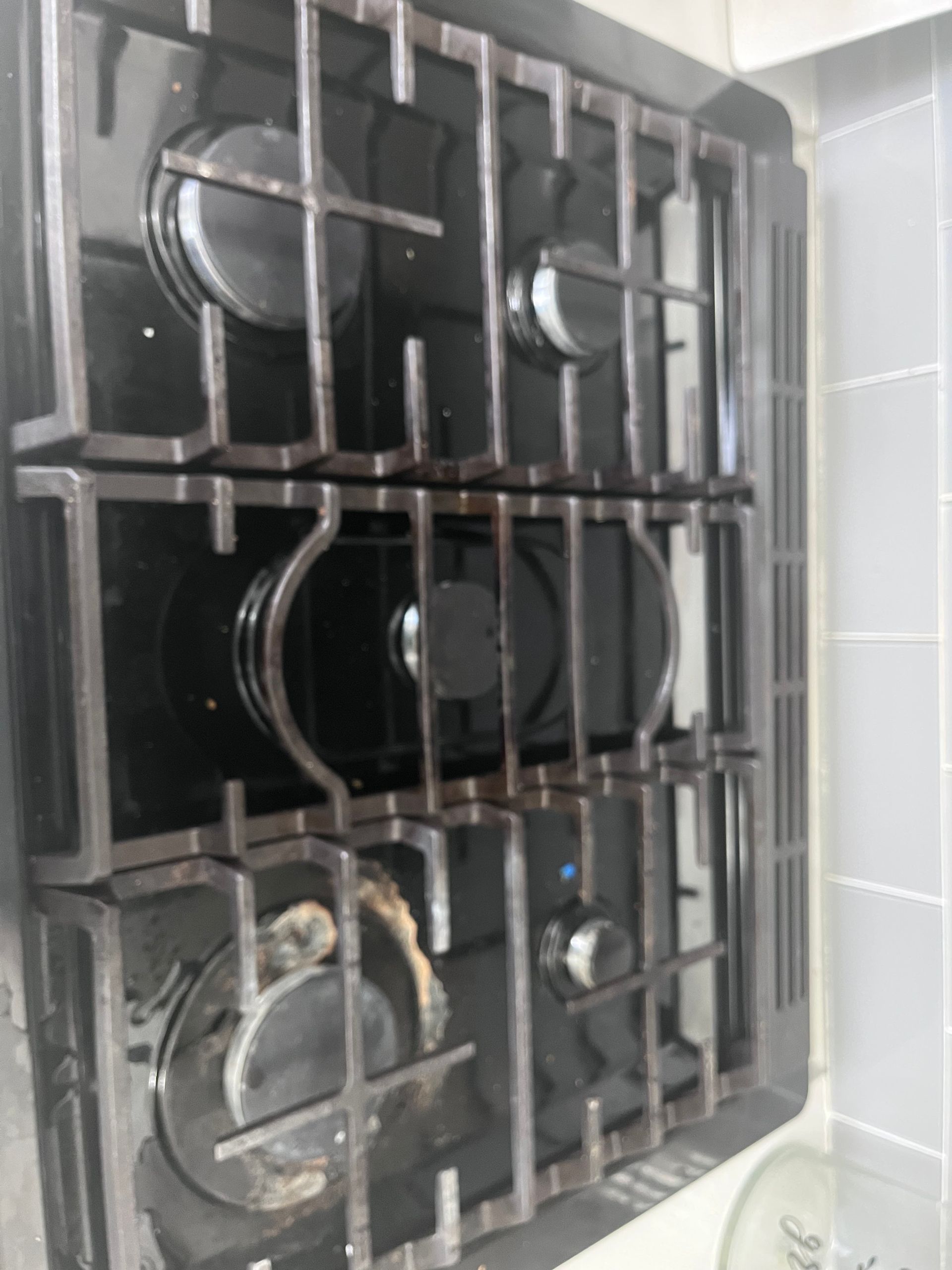 A black gas stovetop with four burners and grates, showing some dirt and food residue. It's set into a white tiled counter.