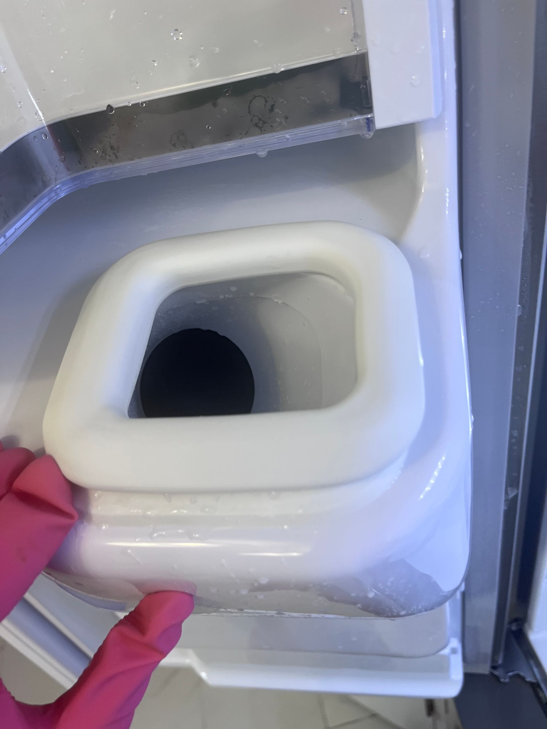 A person wearing a pink glove holds a white ice bin's handle in a refrigerator. Ice crystals are visible.