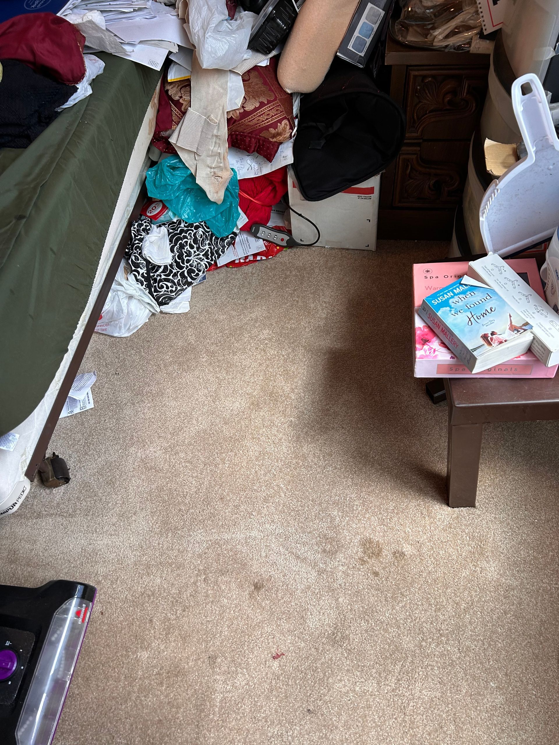 Messy room with scattered clothes and objects. A small table holds a book and remote.