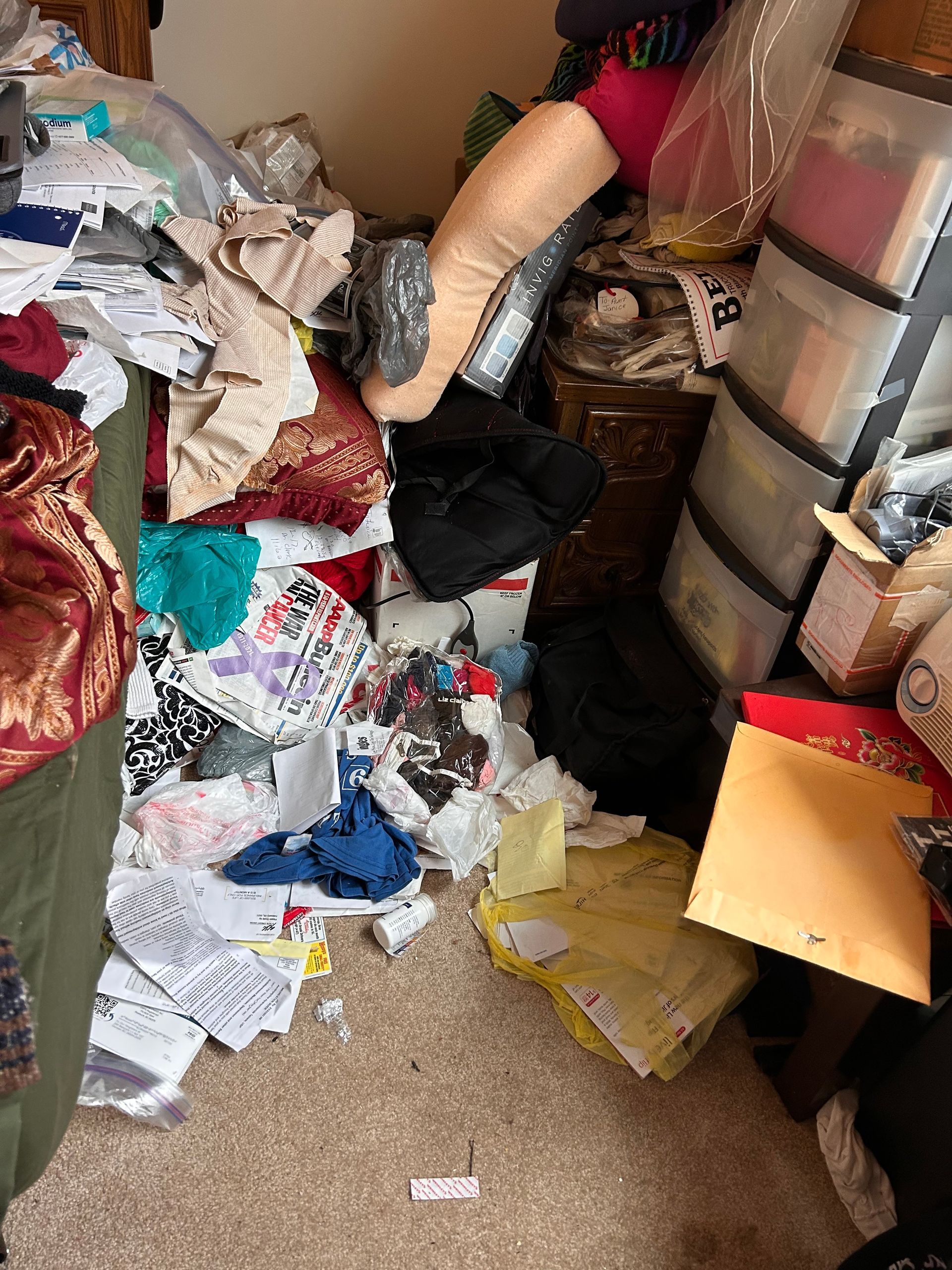 A cluttered room with piles of fabrics, papers, and containers; a person's leg is visible amongst the mess.