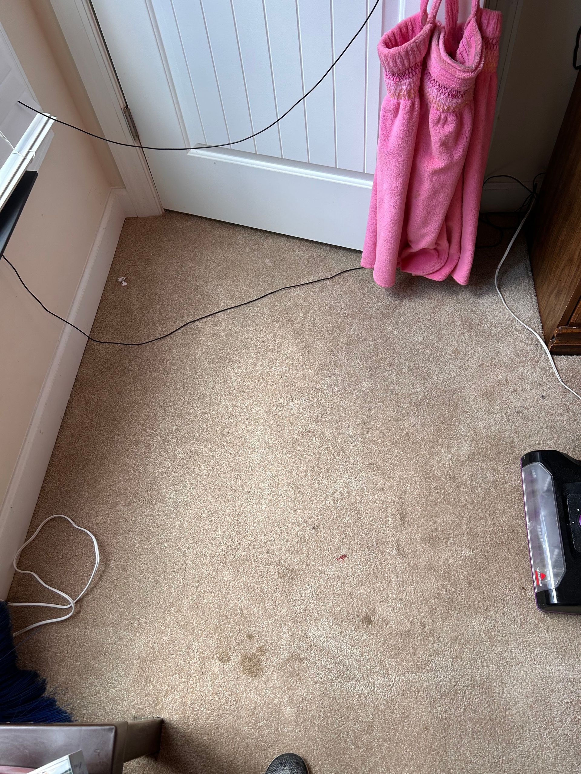 Beige carpet with visible stains and cords. A pink dress hangs on a white door. A vacuum cleaner is partially visible.