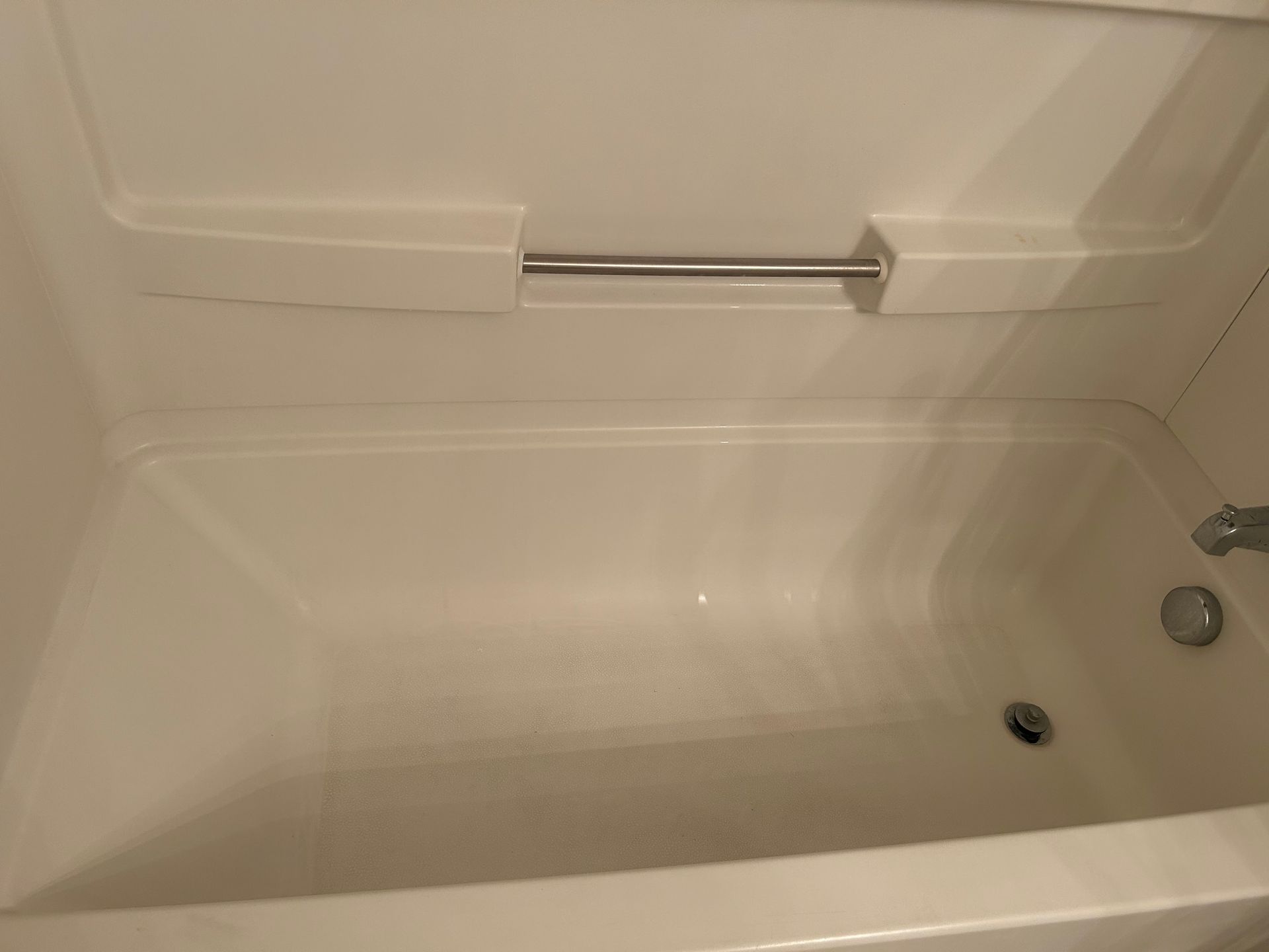 White bathtub with a chrome handrail, viewed from inside the tub. The tub and surrounding wall are clean and empty.