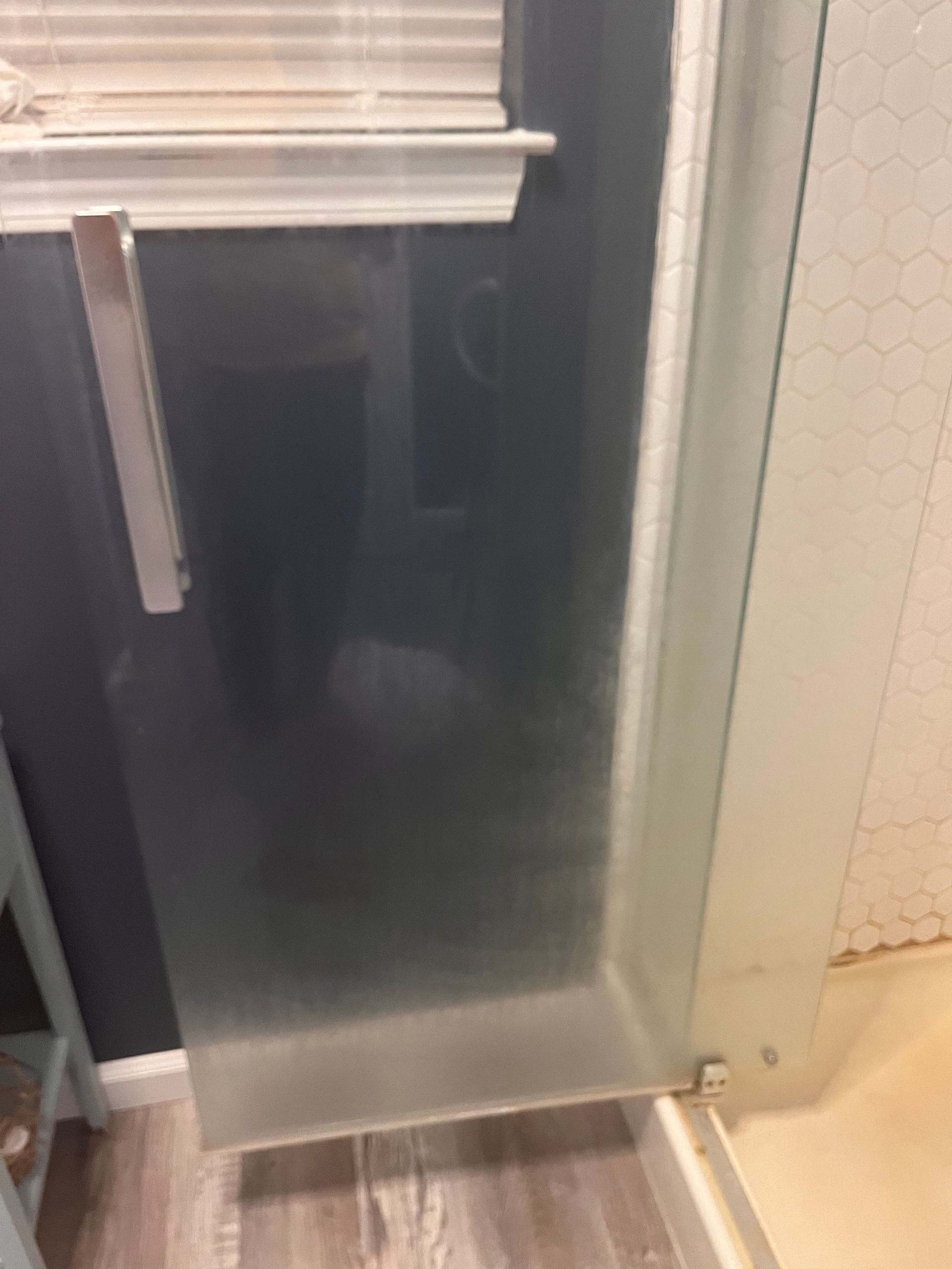 Shower door ajar, showing frosted glass and frame. In the background, a partial view of a window and a tiled wall.