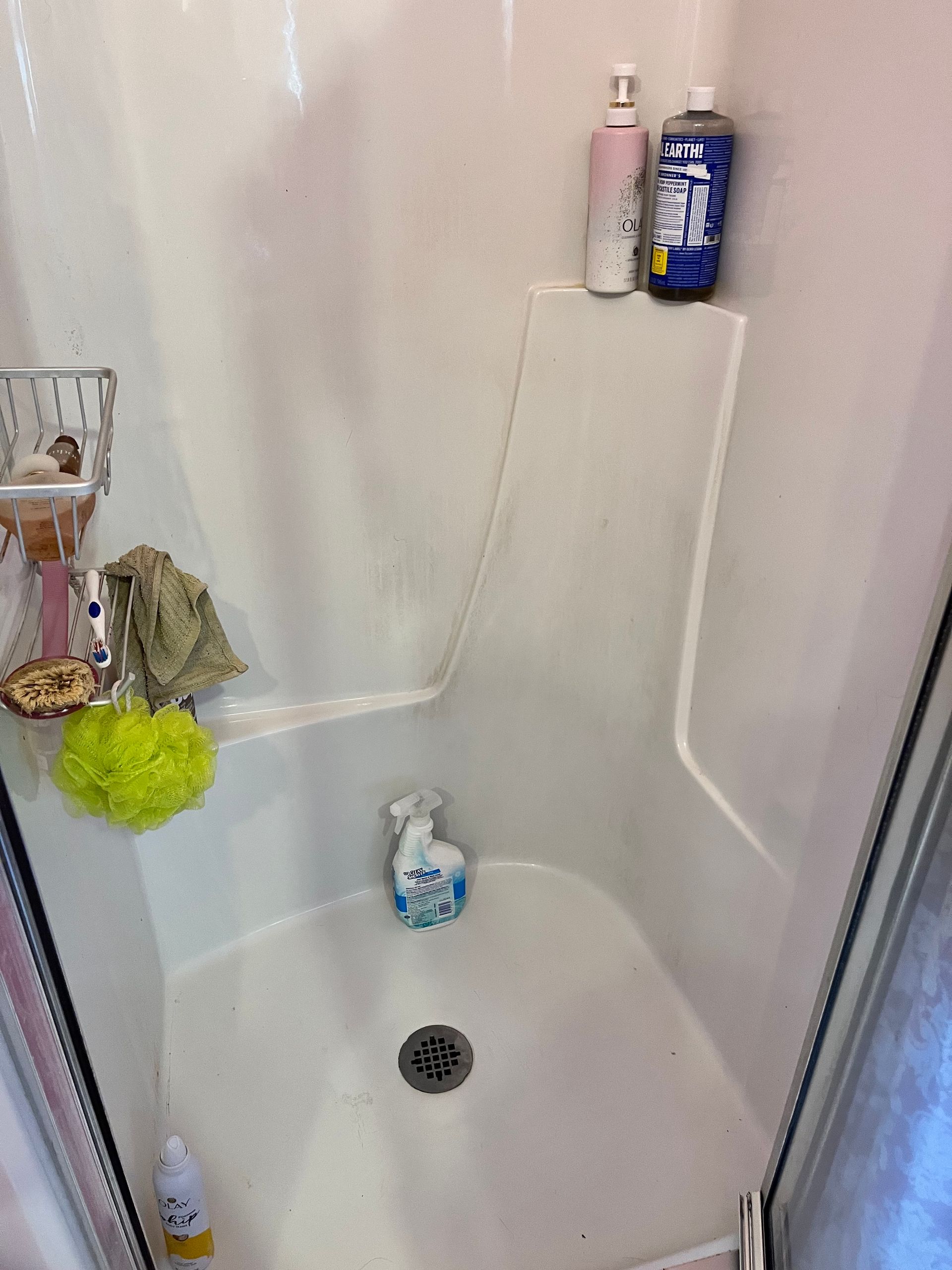 A white shower with soap bottles on a shelf and a metal drain. A loofah and small shelf with toiletries are on the left.