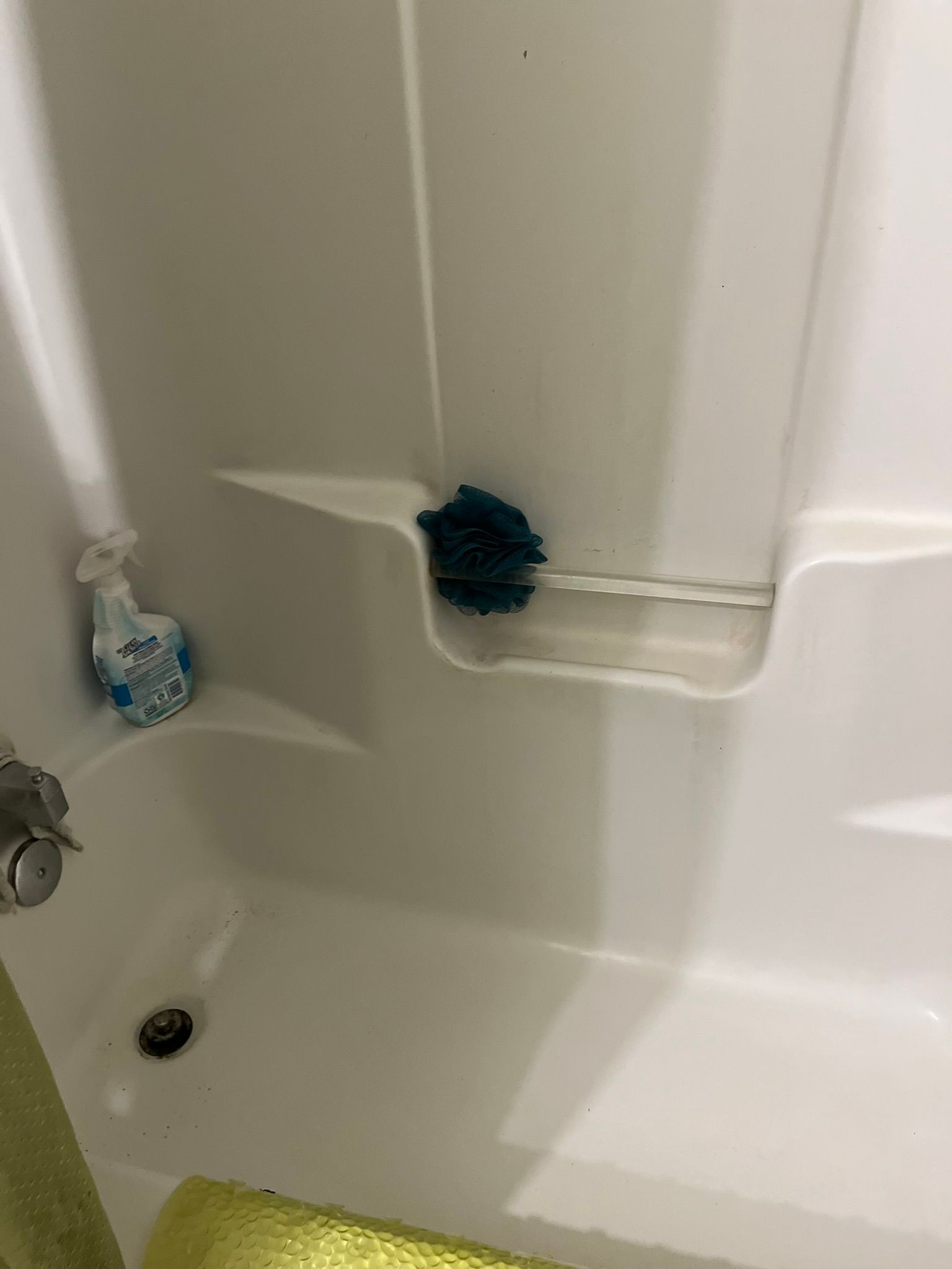 A white bathtub with a teal loofah and cleaning supplies on a shelf in the corner.