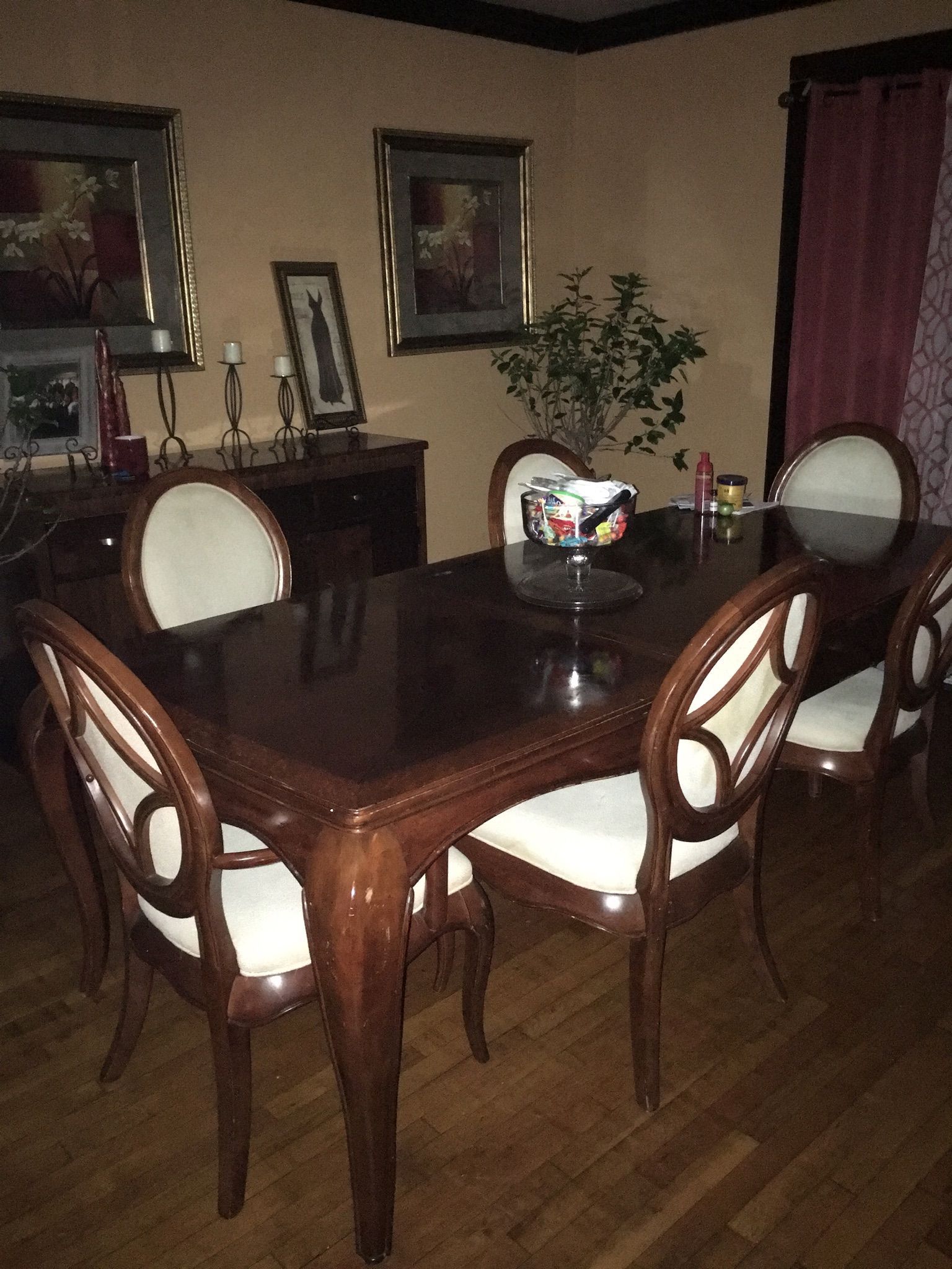 Dining room with a dark wood table, white-cushioned chairs, and artwork on the wall. A plant and candles are visible.