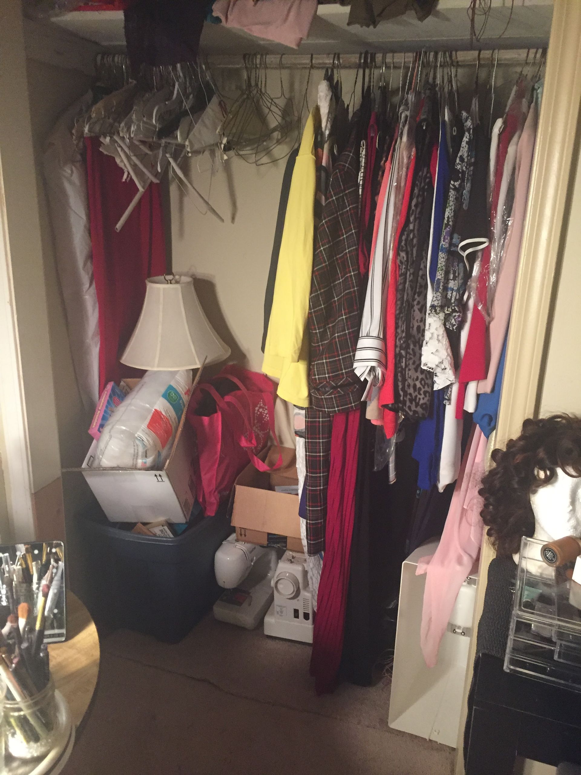 A cluttered closet filled with clothes, a lamp, and storage, with a partially visible makeup station in front.
