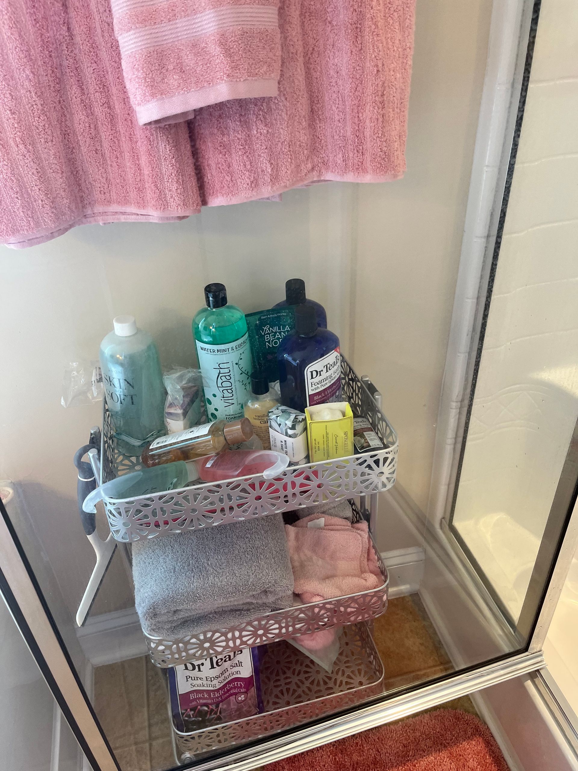 A metal cart in a shower stall holds toiletries, towels, and other bath items. A pink towel hangs above.