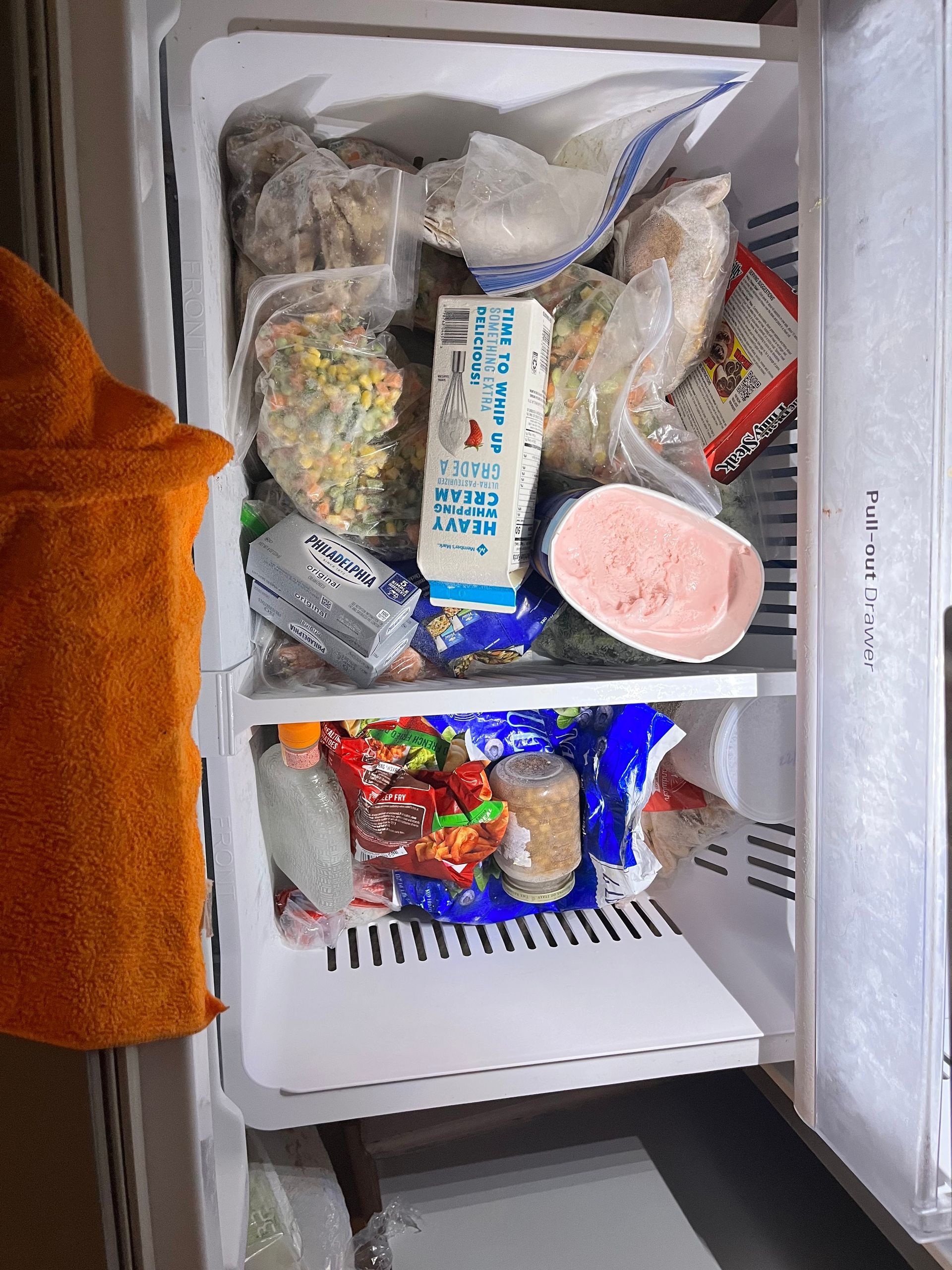 Open freezer filled with frozen food items, including bags, containers, and cartons of milk. Orange towel hangs on the side.