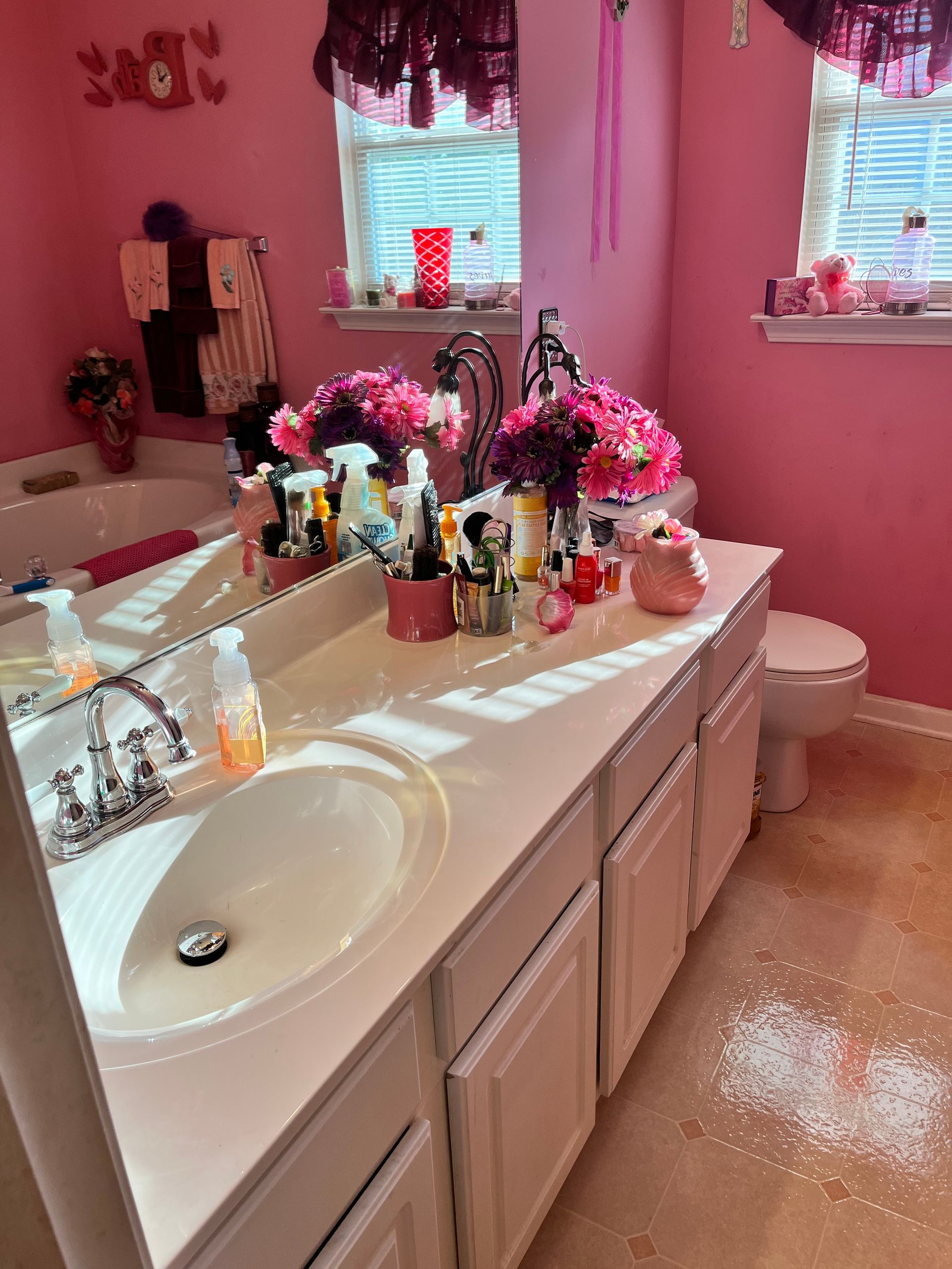 Pink bathroom with a double vanity cluttered with makeup and flowers. A toilet is visible,