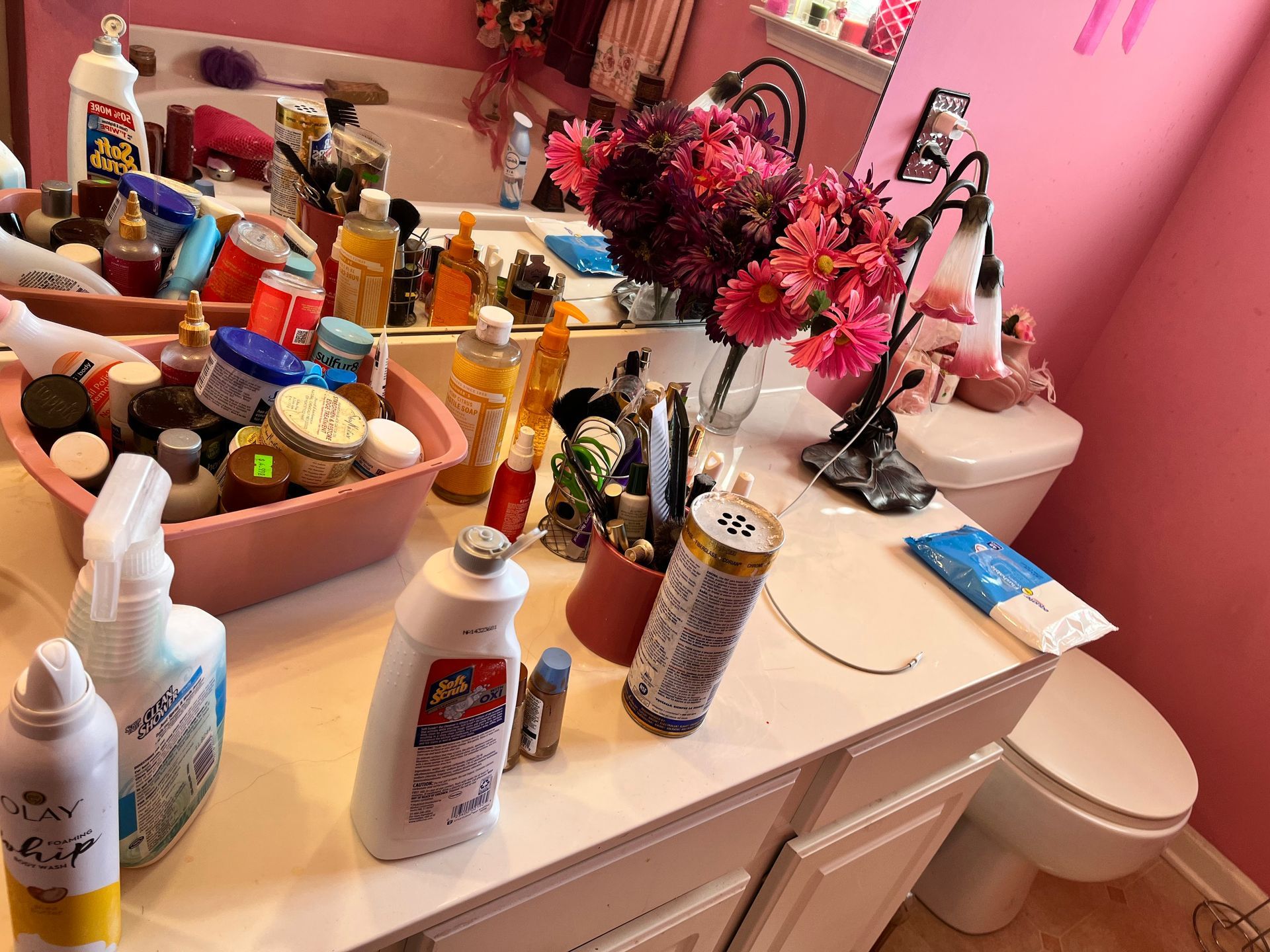 A cluttered pink bathroom vanity with beauty products, flowers, and a toilet.