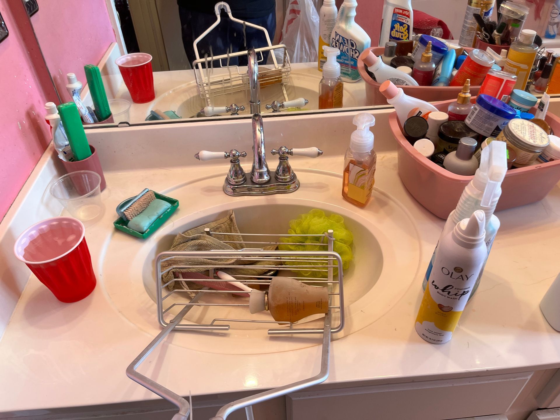 A cluttered bathroom sink with cleaning supplies, cups, and toiletries. A metal rack holds soap and a sponge.