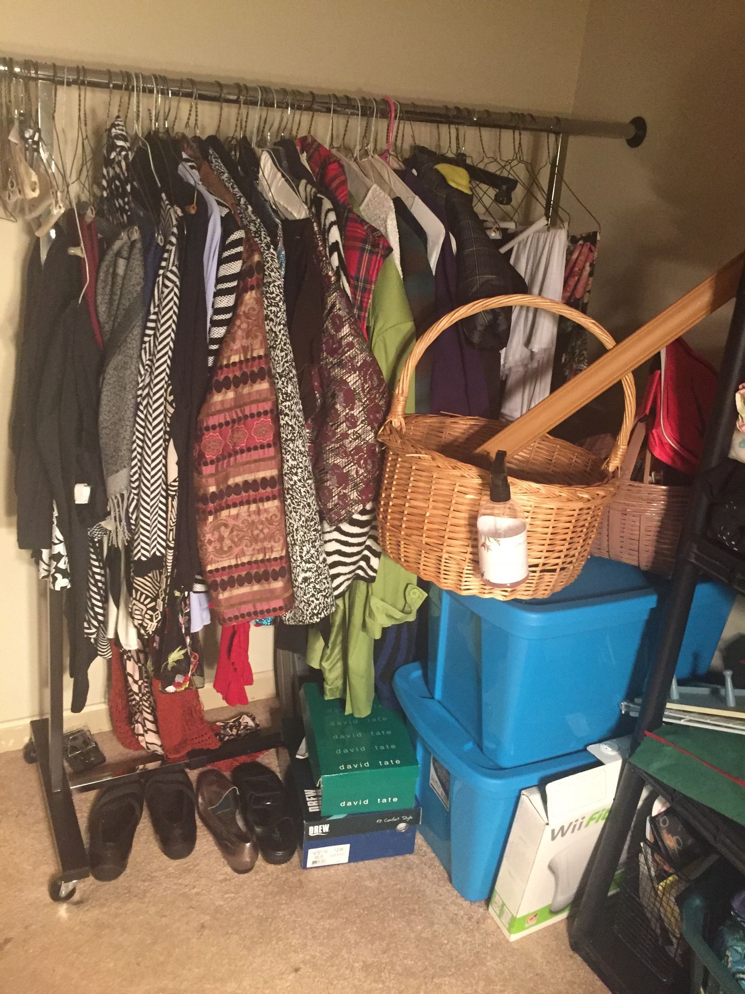 A cluttered clothing rack overflowing with clothes, shoes, and baskets. Storage bins and a cane are nearby in a home setting.