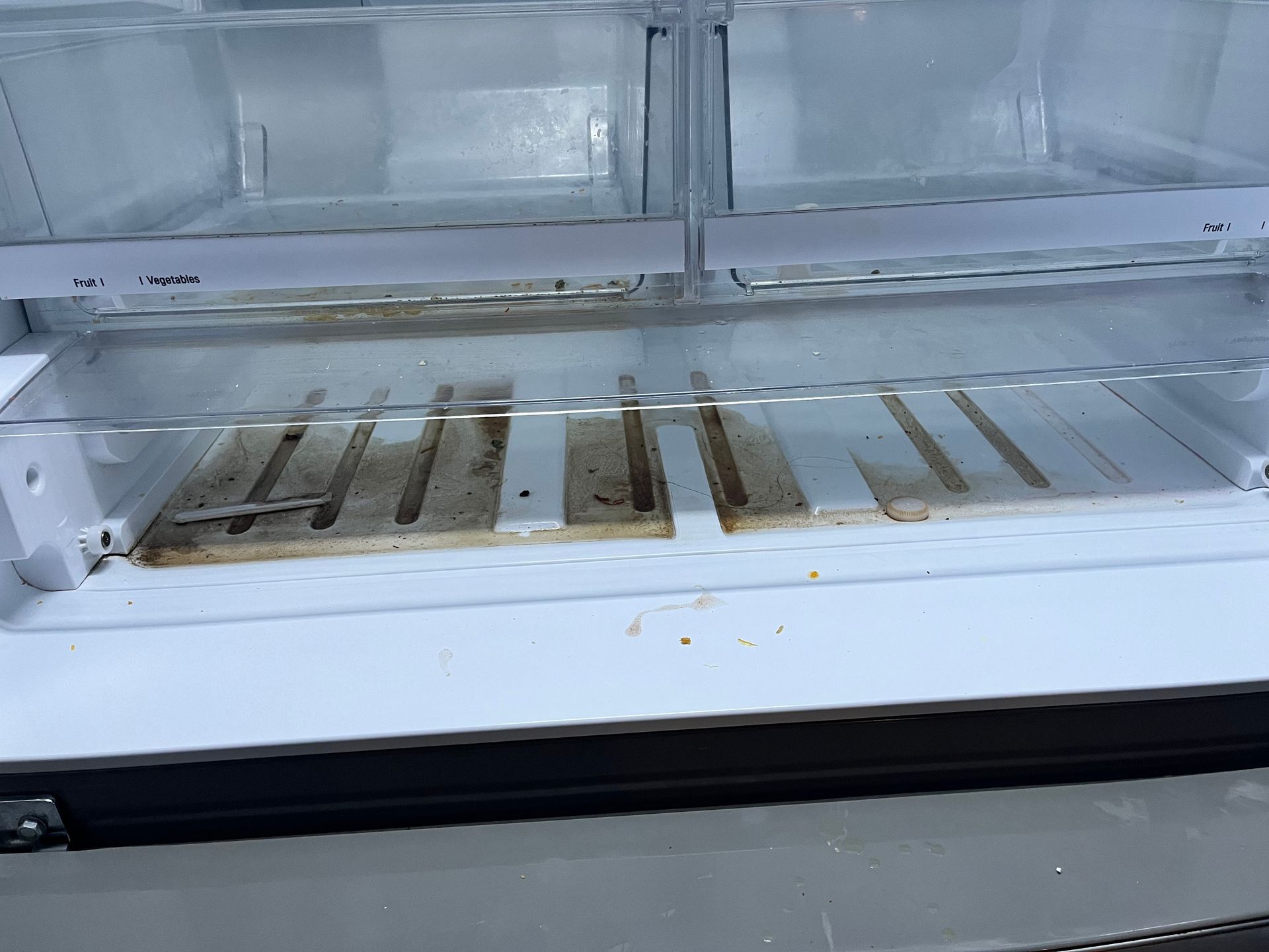 Empty refrigerator, inside view, with dirty food debris on the bottom shelf and door compartments.