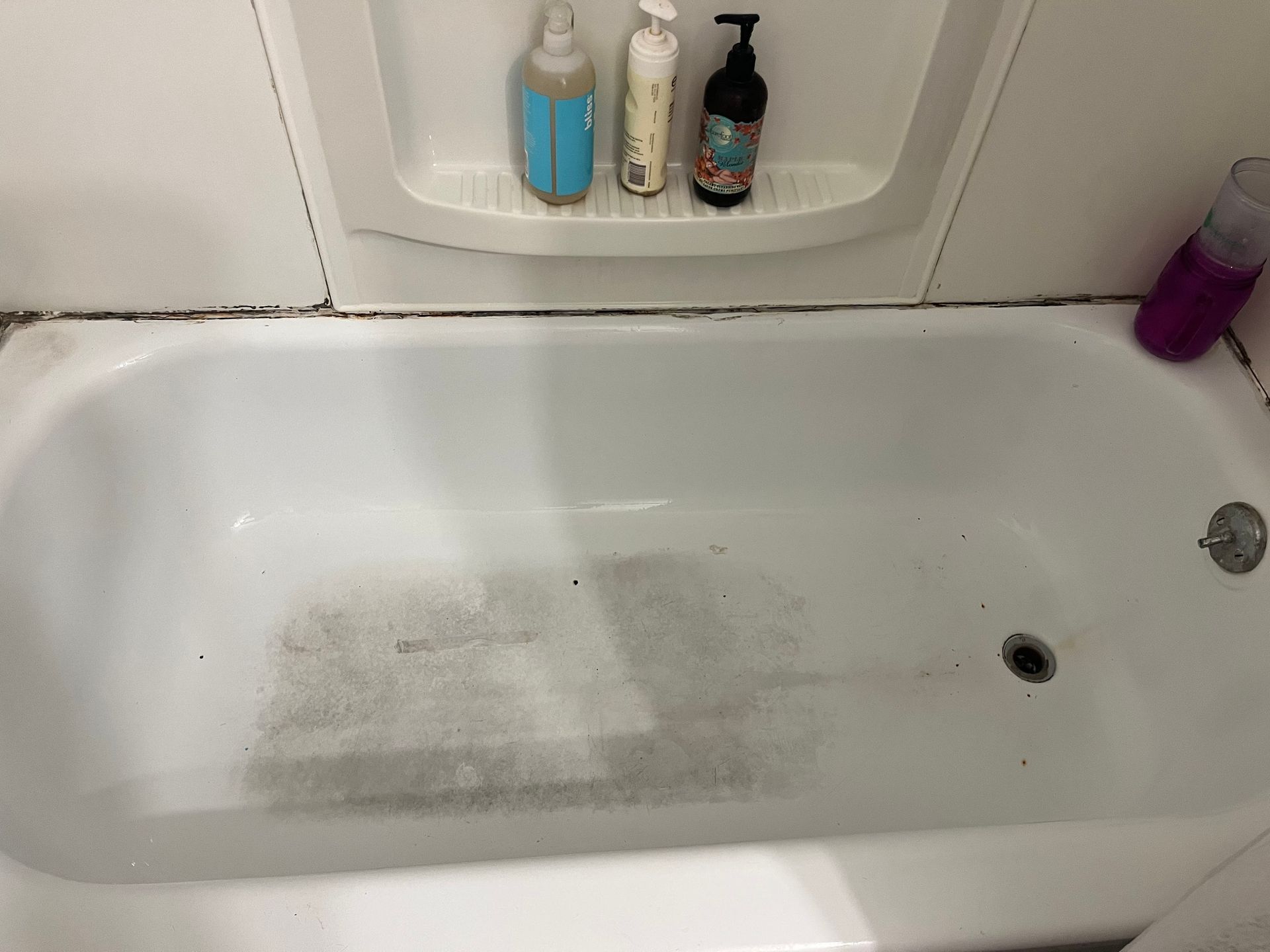 A dirty white bathtub with visible stains and mold around the edges. Shelves above hold toiletries.