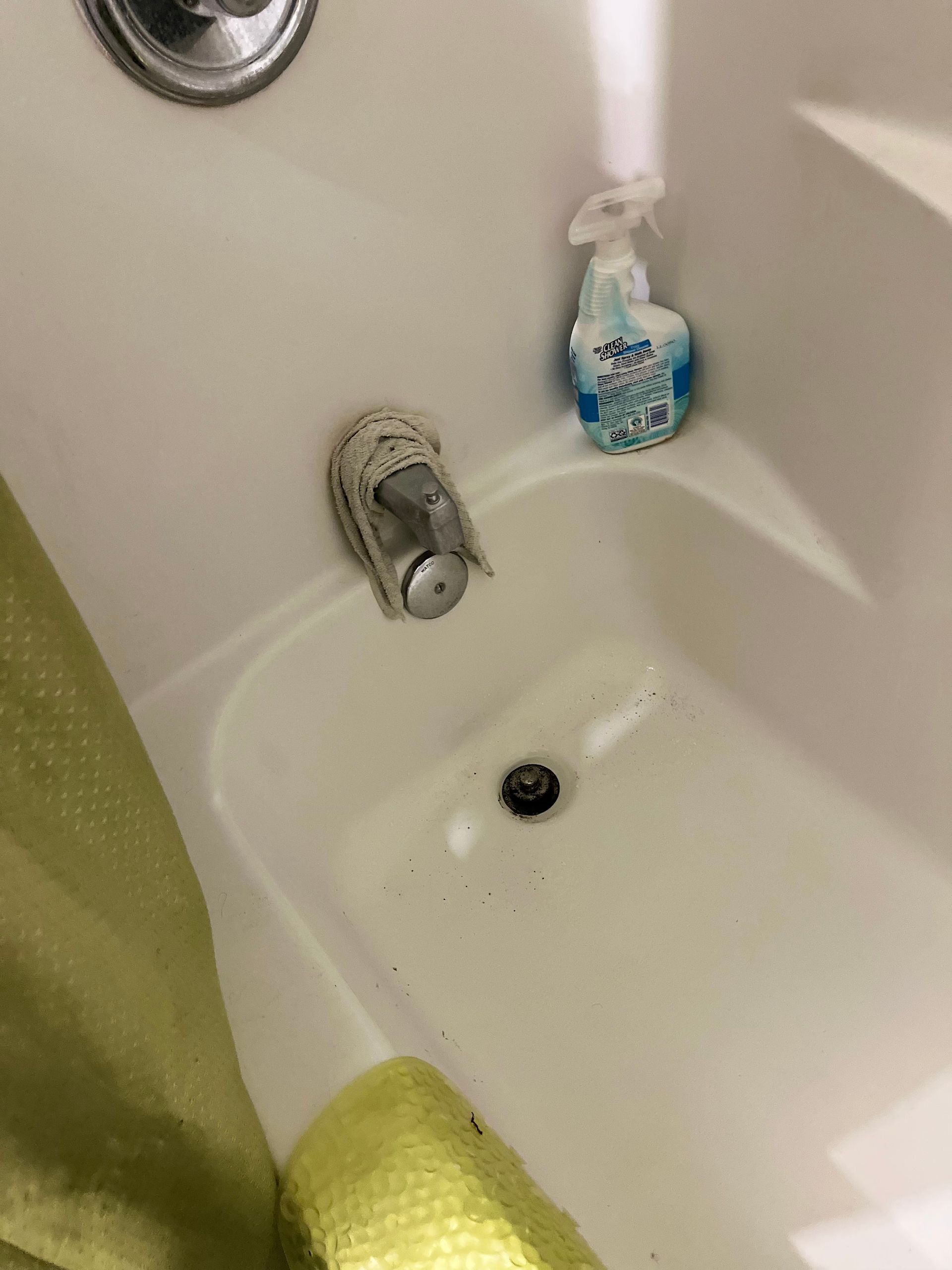 White bathtub with fixtures, a cleaning spray bottle, and a glimpse of a yellow towel and green shower curtain.