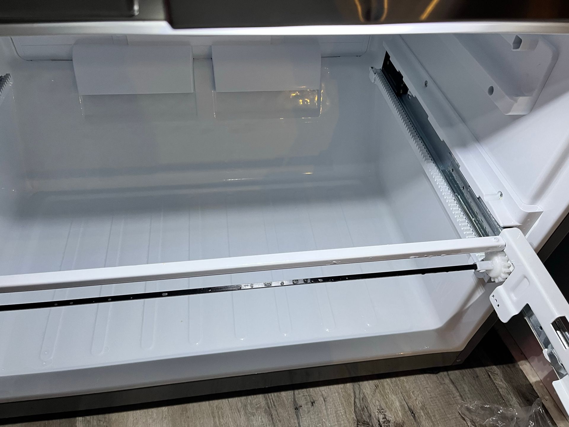 Inside view of an empty, white freezer drawer in a refrigerator, showing the drawer's interior and sliding tracks.