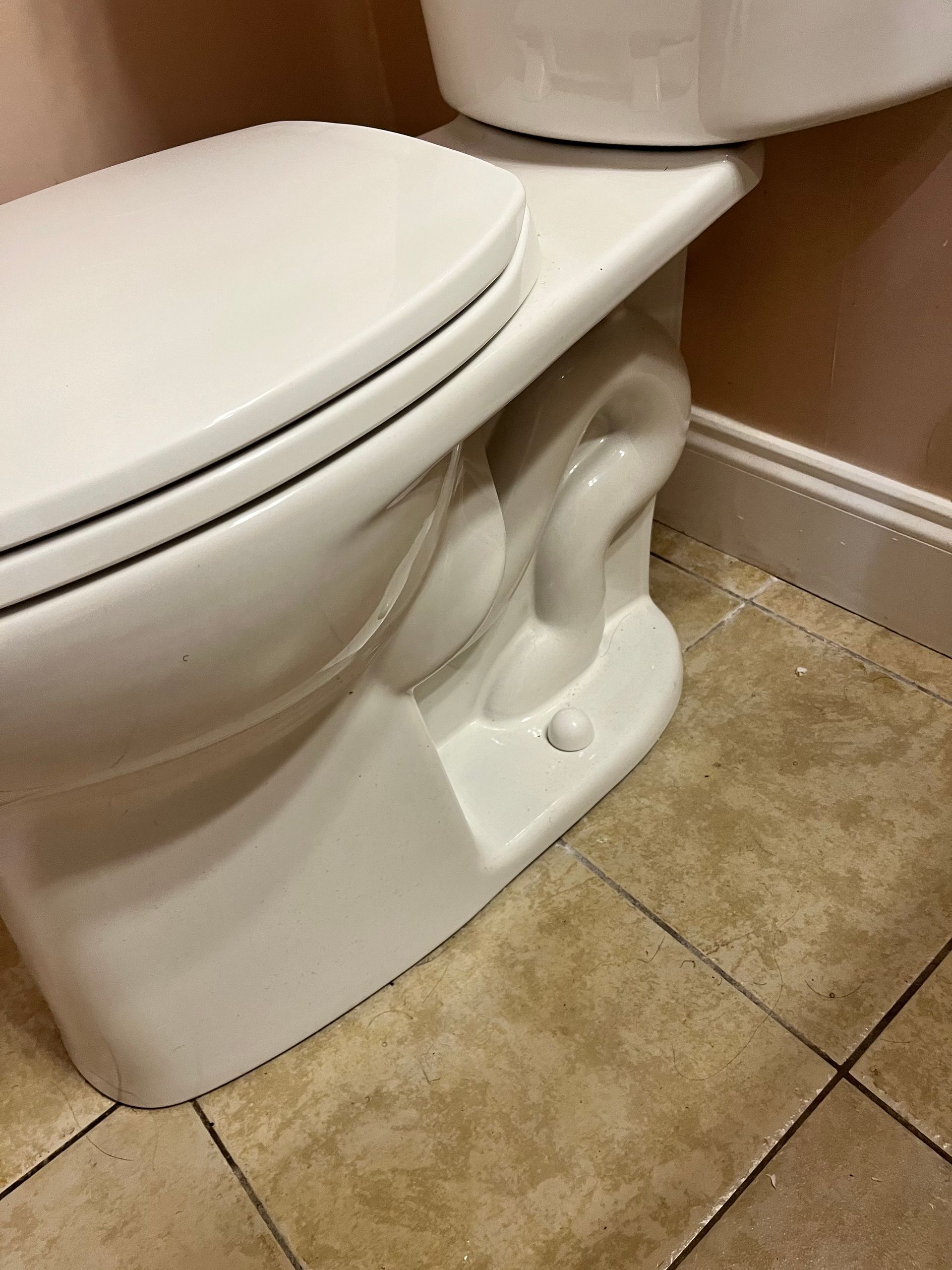 White toilet with a closed lid, set against a pink wall and tan tile floor. The toilet has an ornate, sculpted base.