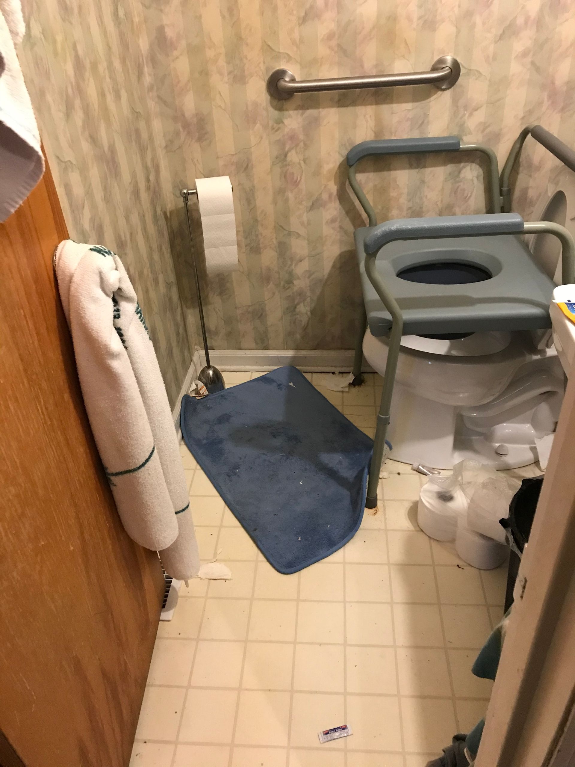 Small bathroom with a toilet, a raised toilet seat, a grab bar, and a small blue rug. A towel hangs on the door.