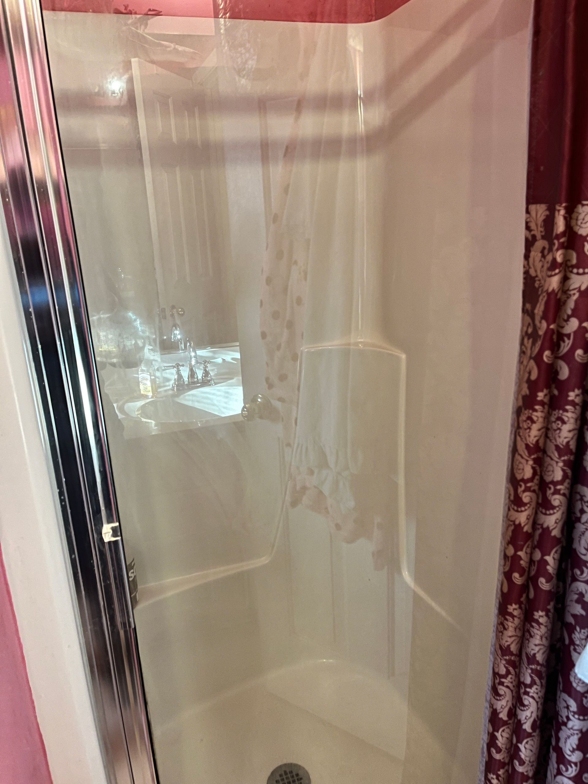 A small, white corner shower with a clear plastic curtain. A partially visible patterned curtain is on the right.