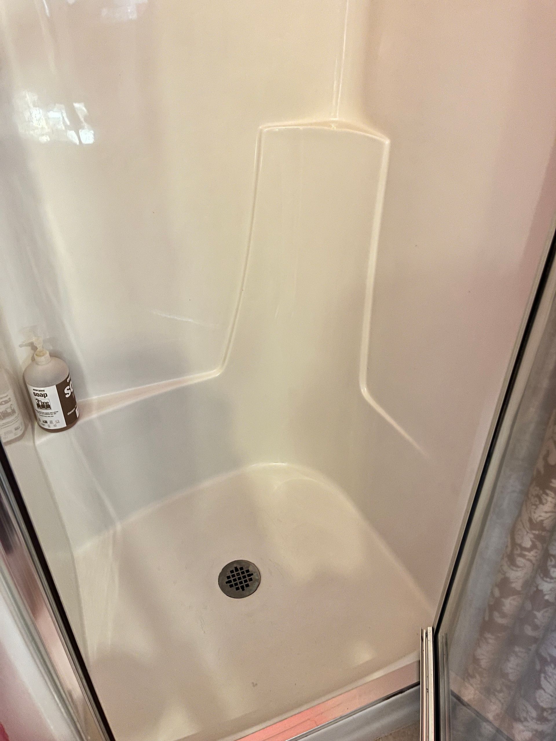White, molded plastic shower stall with a central drain. A bottle of soap sits on a shelf.