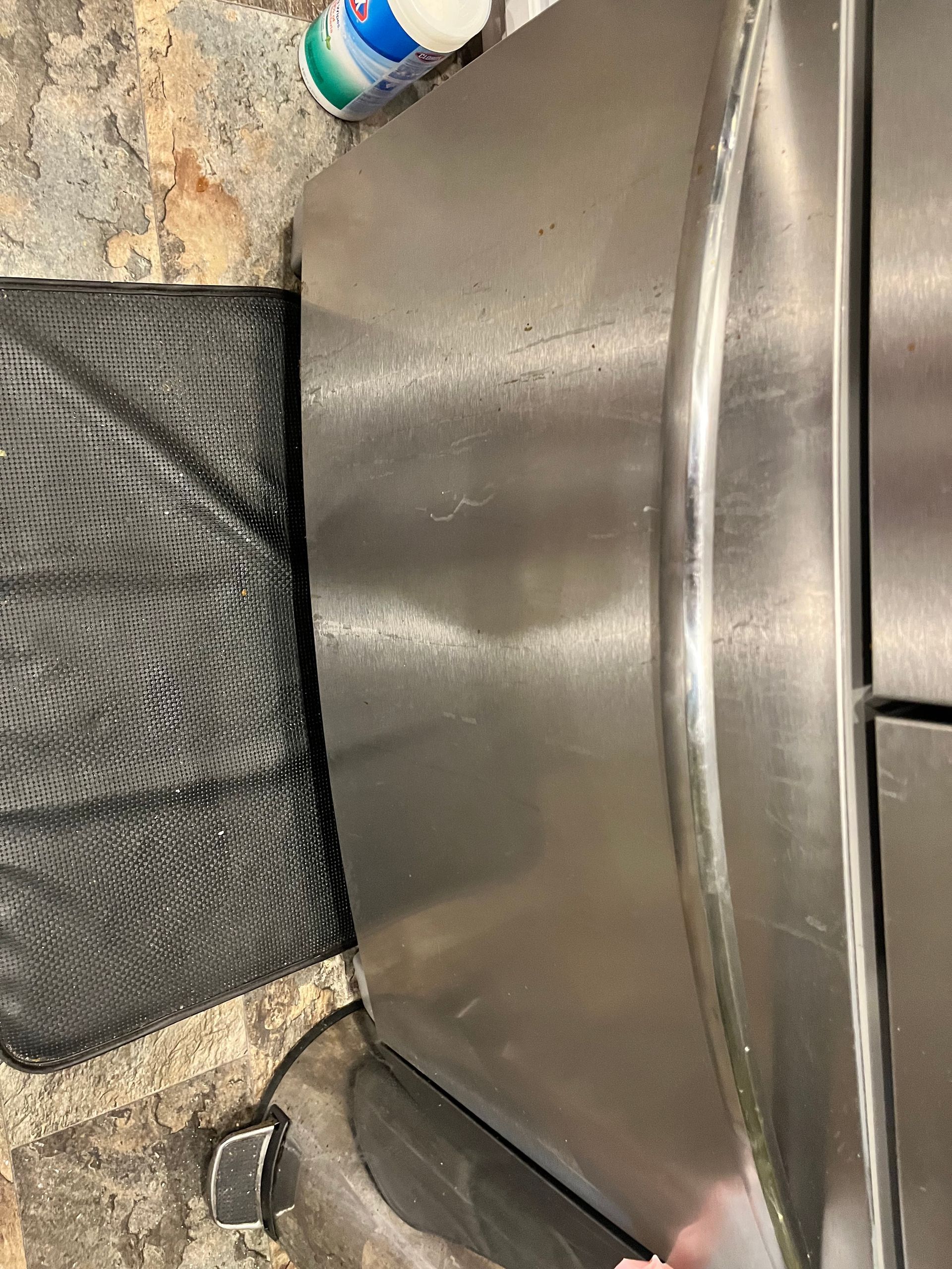 Stainless steel refrigerator door with some streaks and dirt.  A black mat and bottle of cleaner are visible.