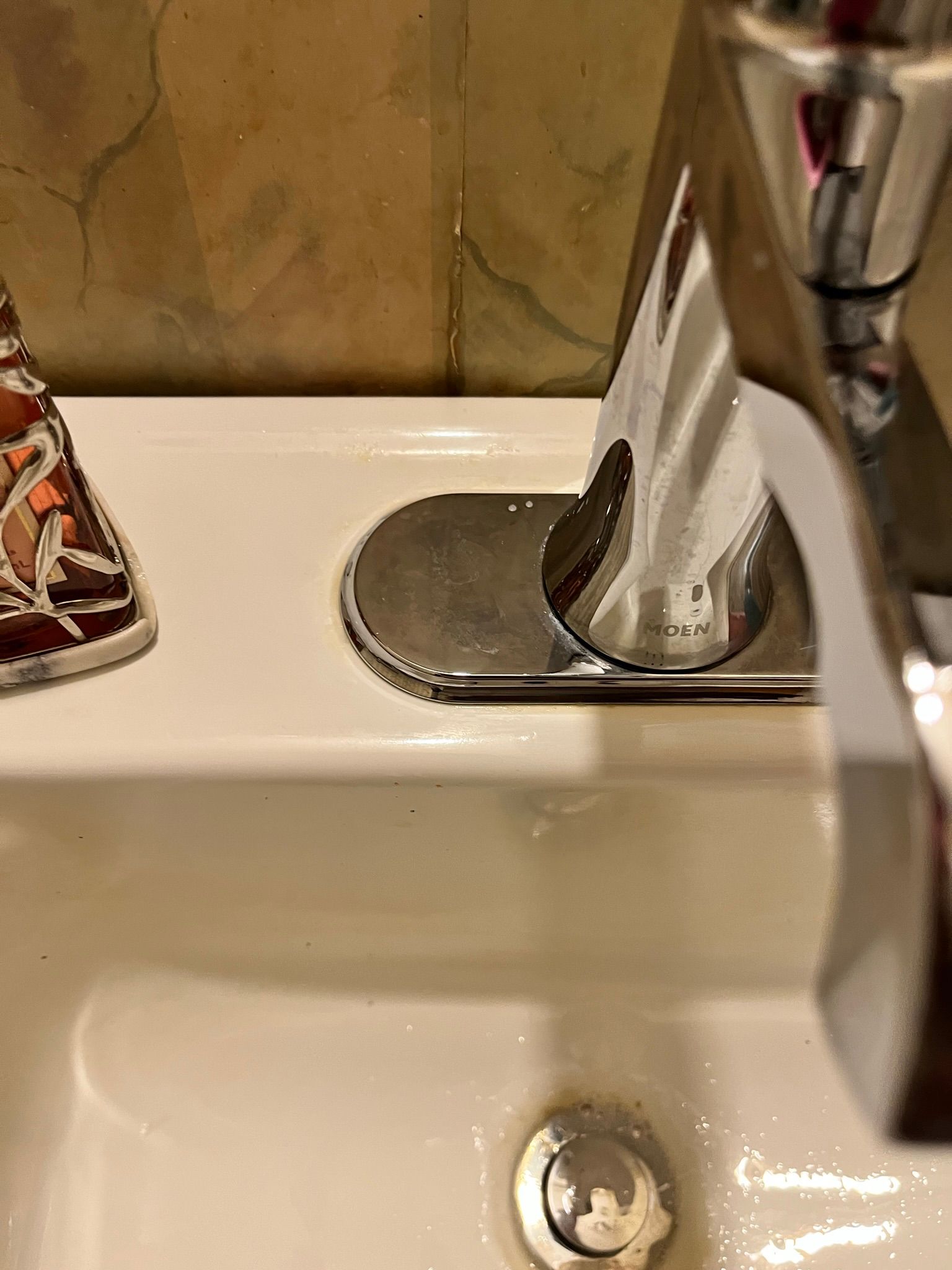 Close-up of a shiny chrome faucet in a white sink. Brown and beige patterned tiles are in the background.