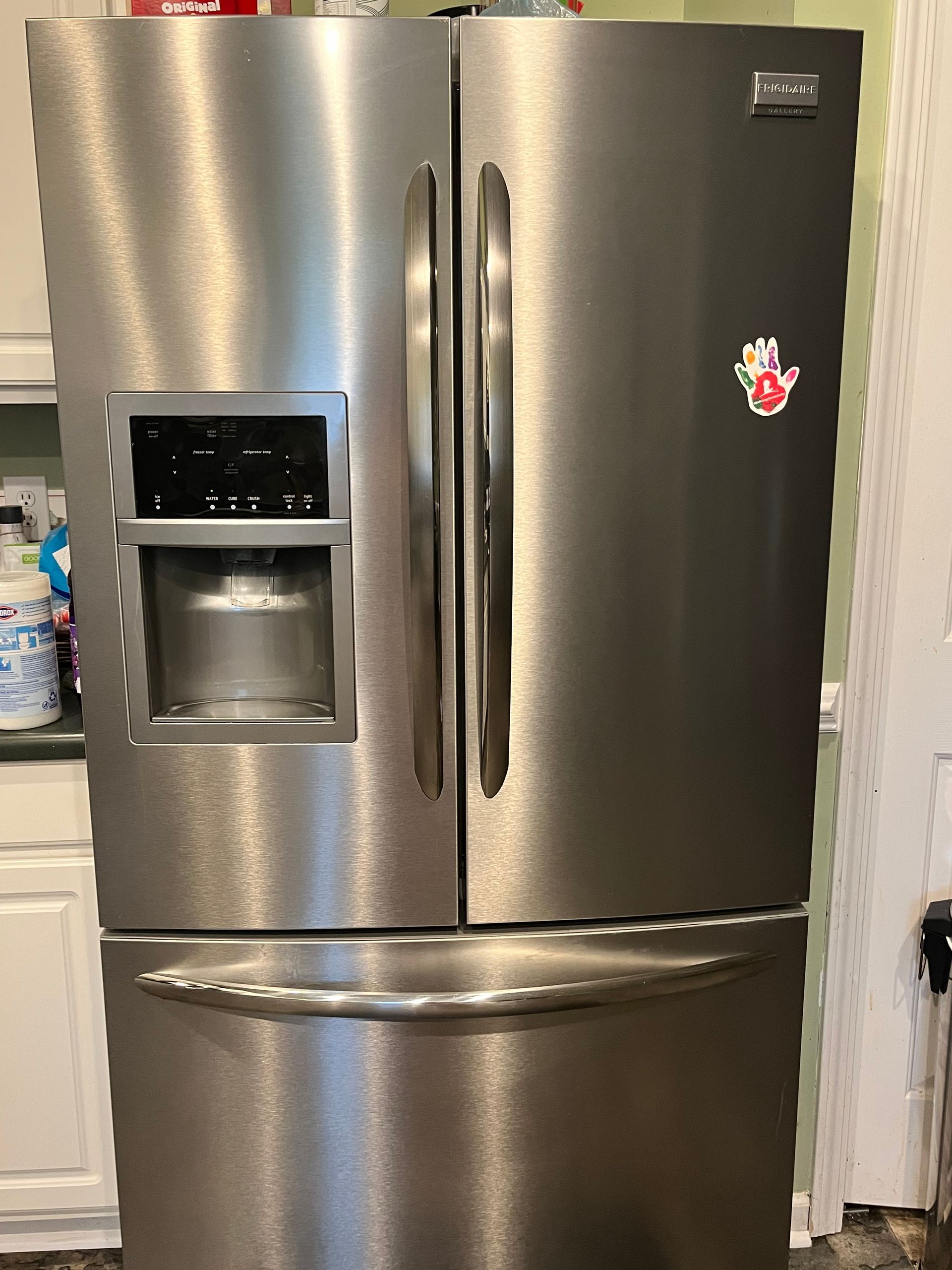 Stainless steel French door refrigerator with water dispenser, in a kitchen setting. Has a sticker on the right door.