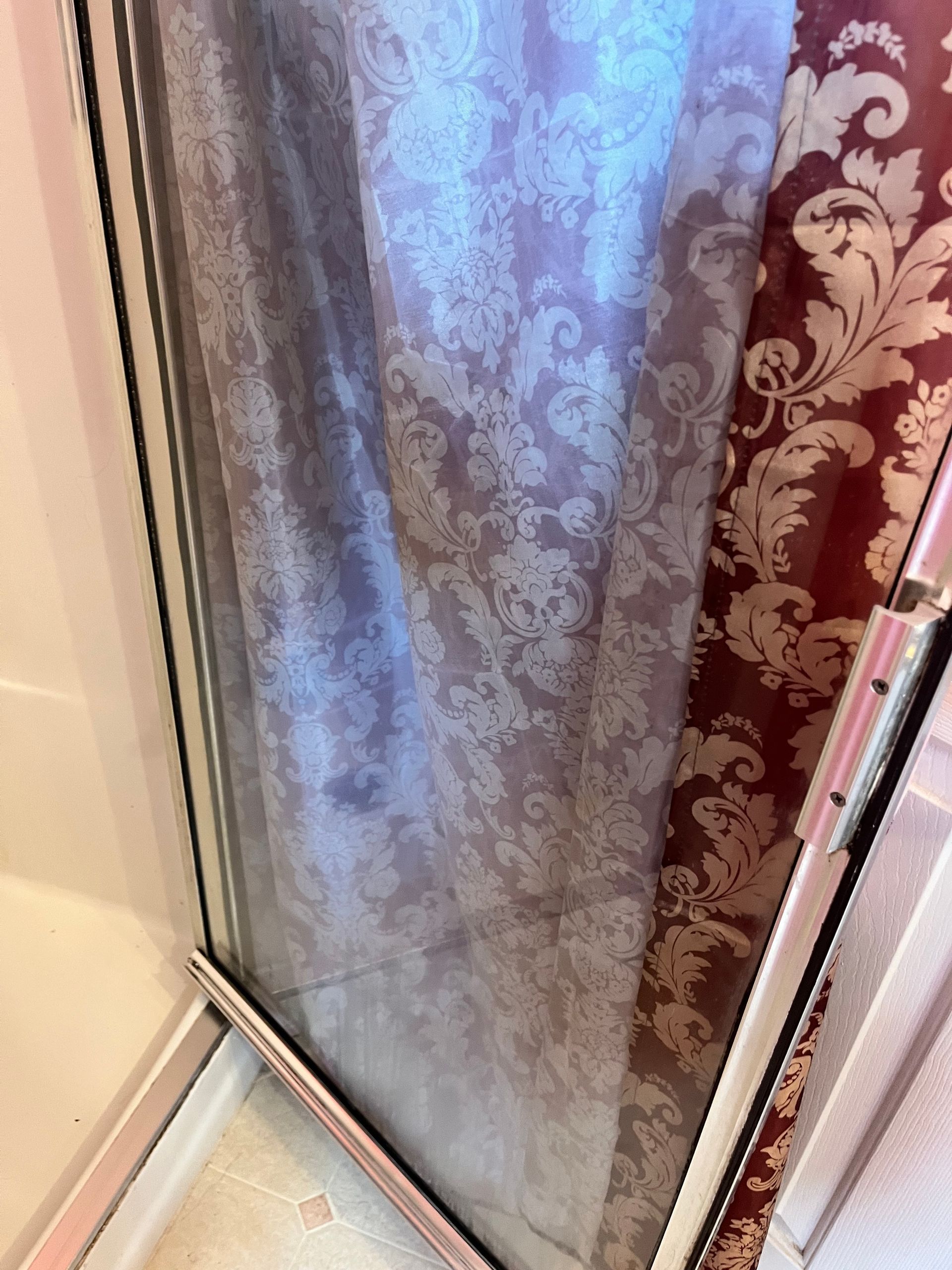 Close-up of window with light purple patterned curtain next to a red floral curtain. The frame is white.