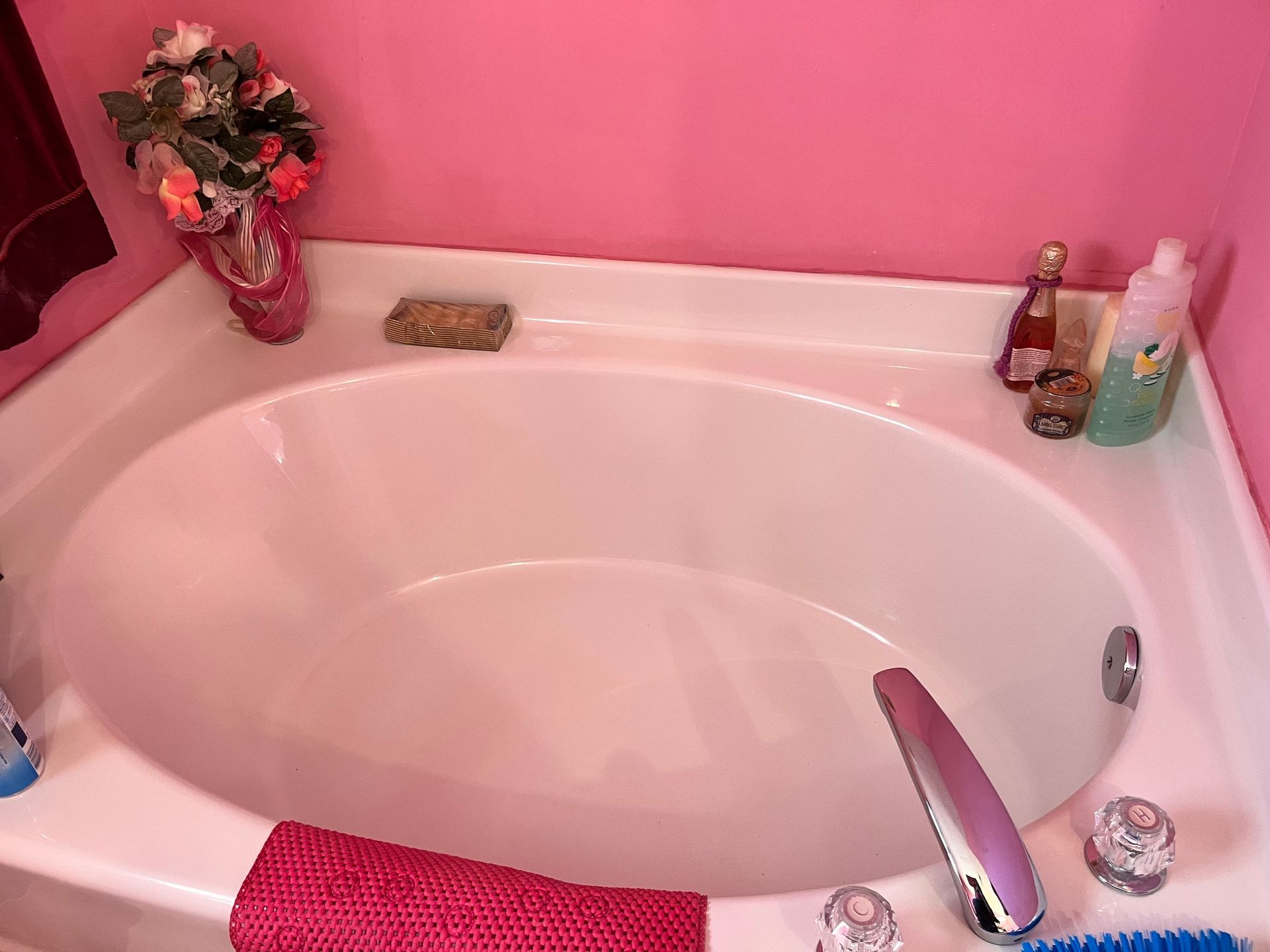 An empty white oval bathtub against a pink wall, with bath products and a towel in the corner.