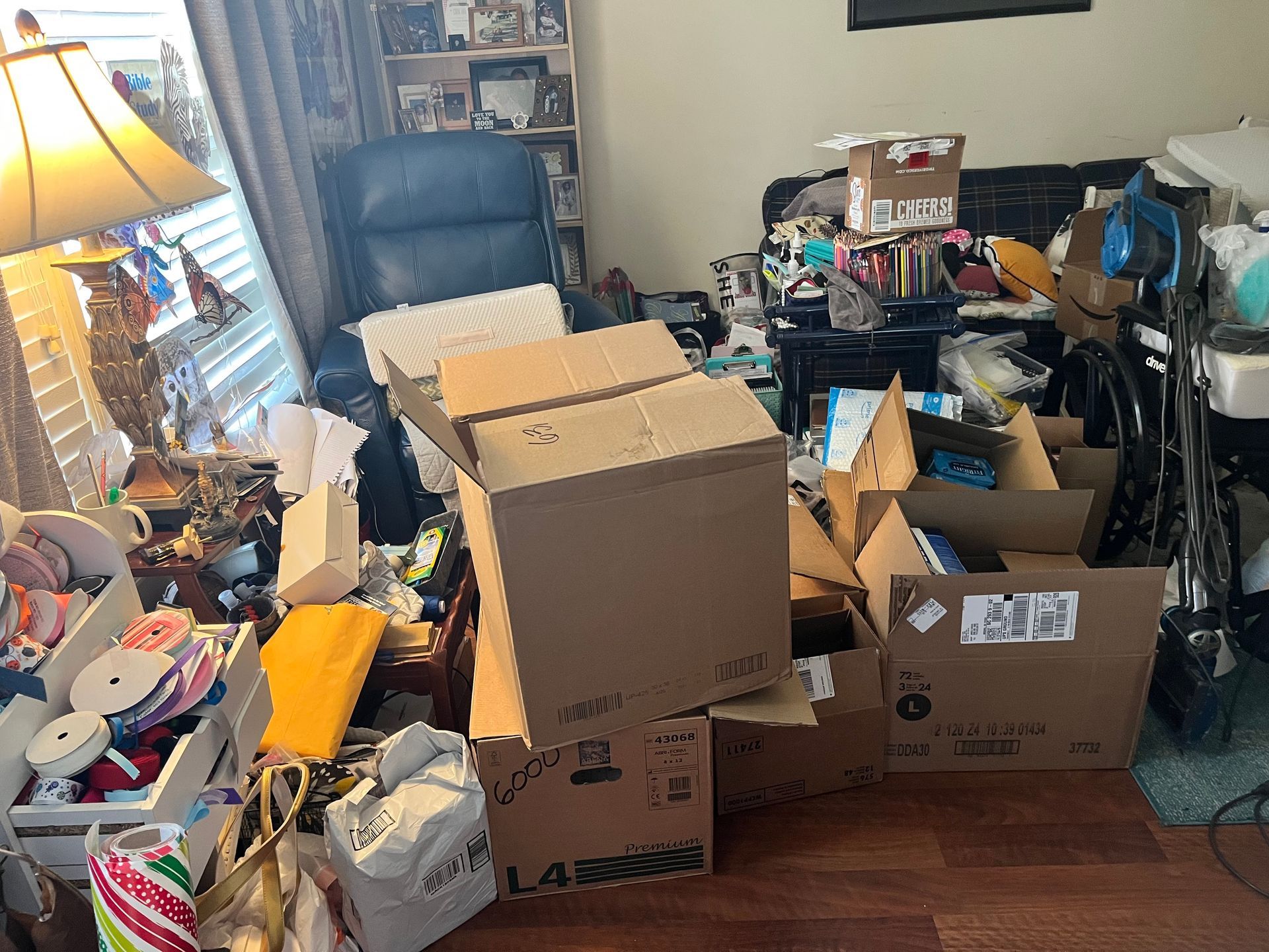 Cluttered living room with boxes and various items scattered around furniture. A lamp sits on a table near a window.