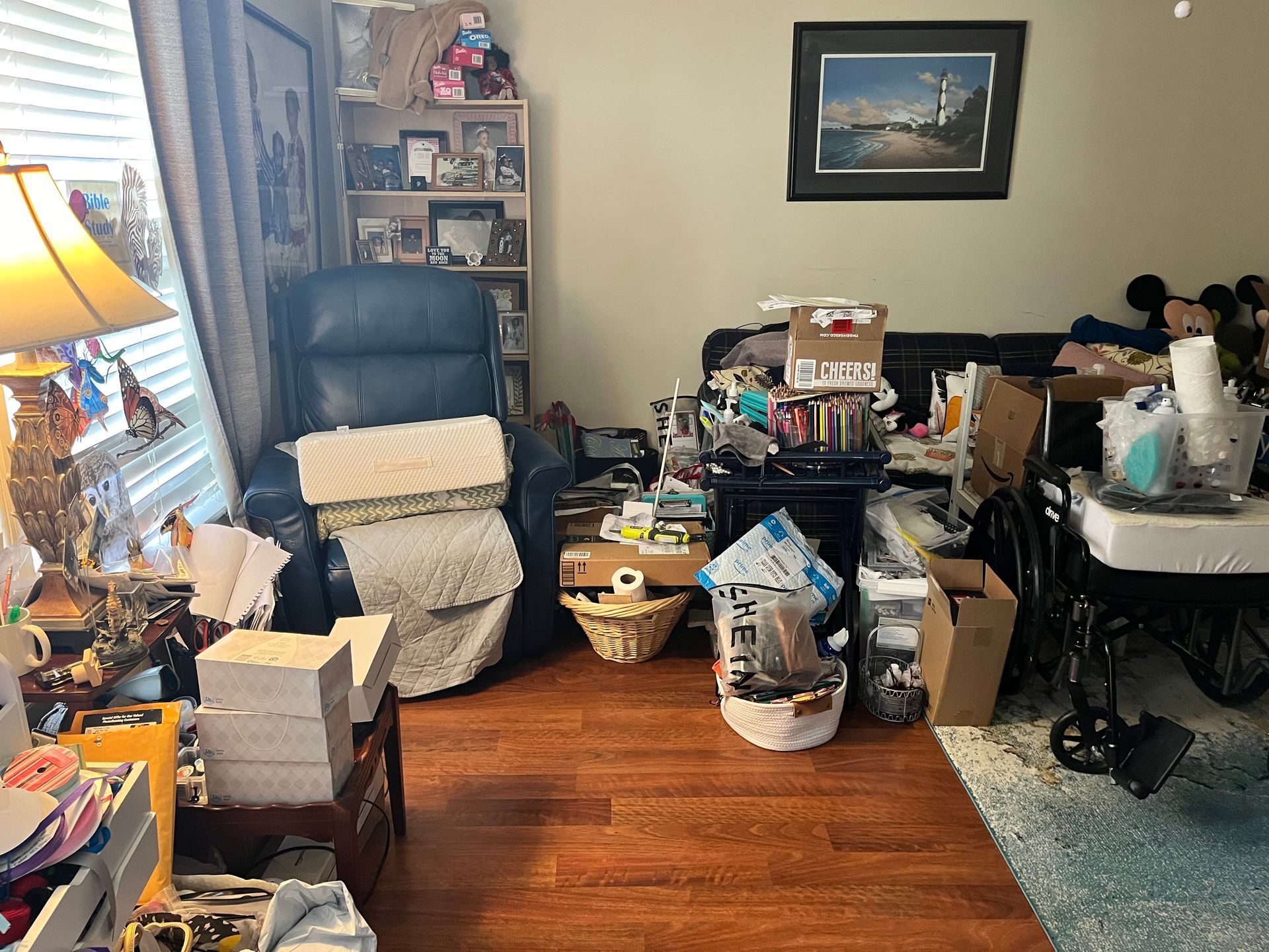 Cluttered living room with a black recliner, boxes, baskets, and various items scattered on the floor and shelves.