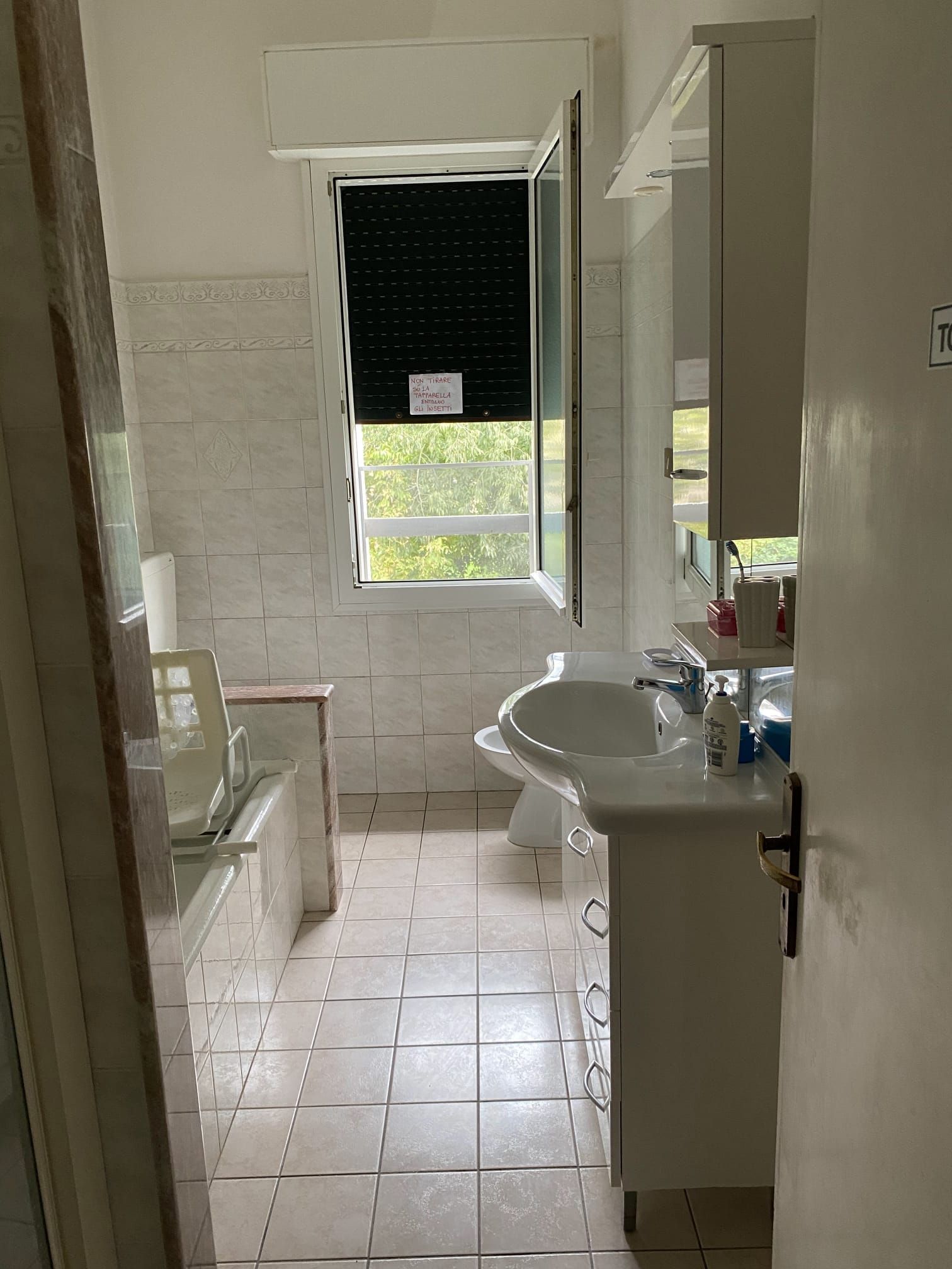 Bagno con sanitari bianchi, finestra con persiana chiusa, pavimento e pareti piastrellati.