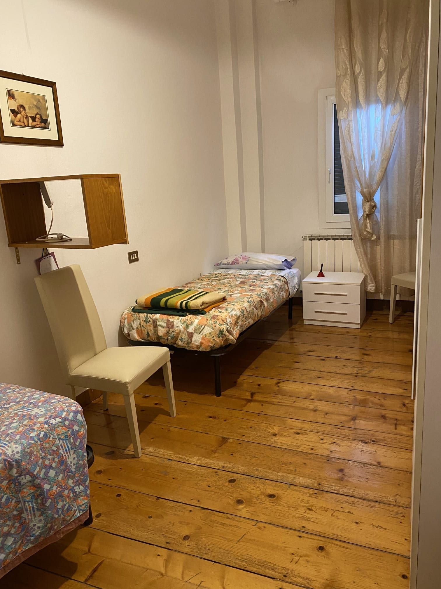 Camera da letto con letto singolo, pavimento in legno, comodino bianco e finestra con tende trasparenti.