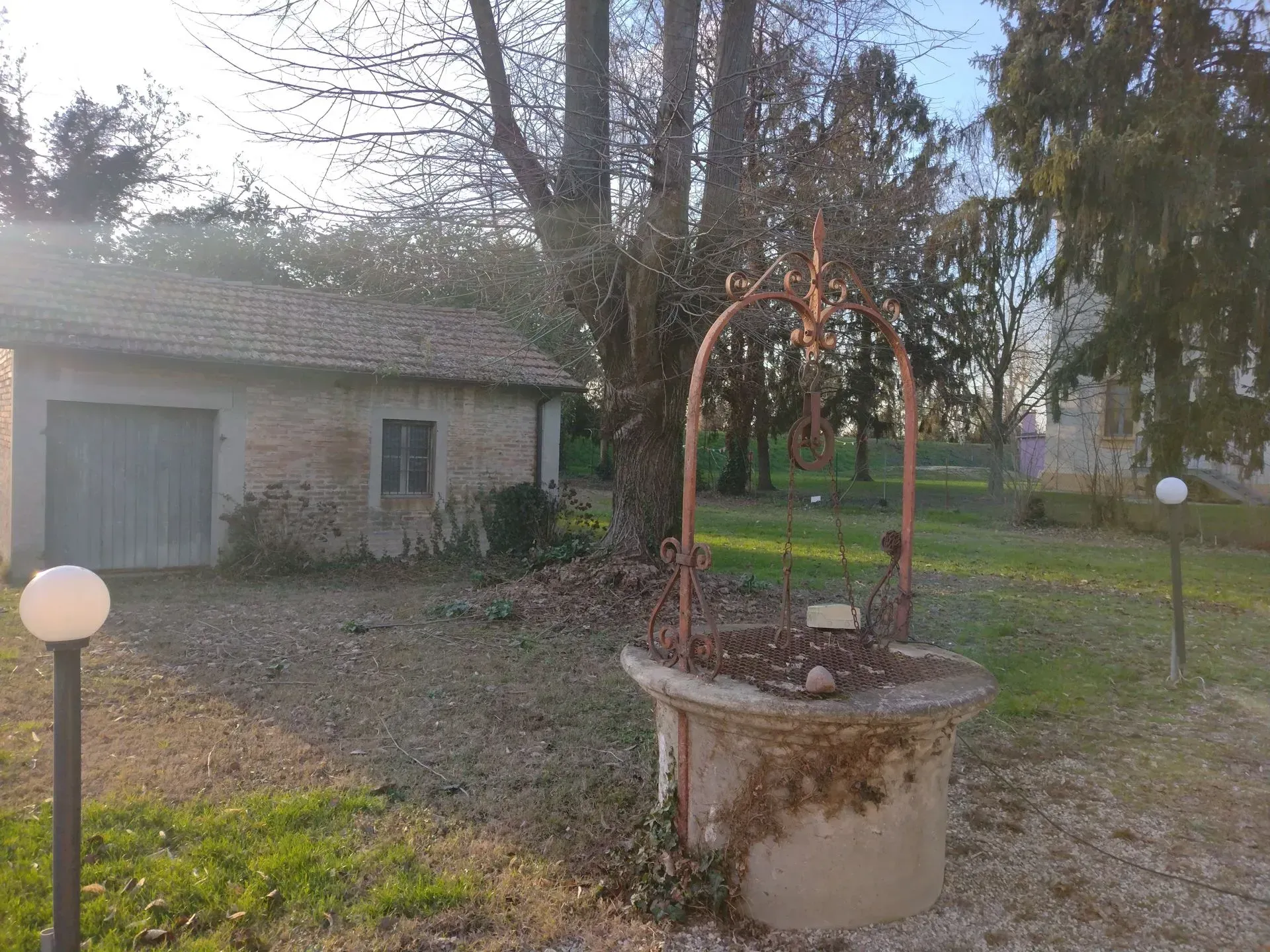 Un vecchio pozzo con un arco di metallo arrugginito, un piccolo edificio con una porta da garage, alberi e due lampade a globo in una zona erbosa.