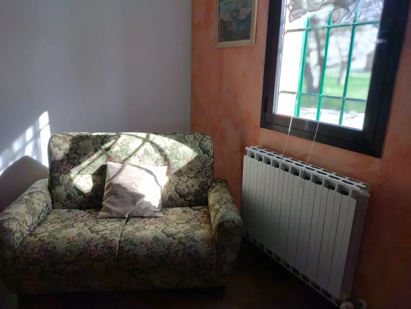 Un piccolo divano floreale con un cuscino è posizionato accanto a una finestra e a un termosifone. La luce del sole filtra a fiotti.