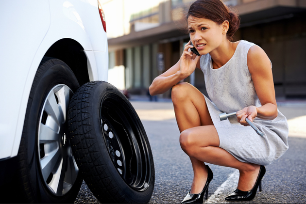 A woman is kneeling next to a tire and talking on a cell phone.