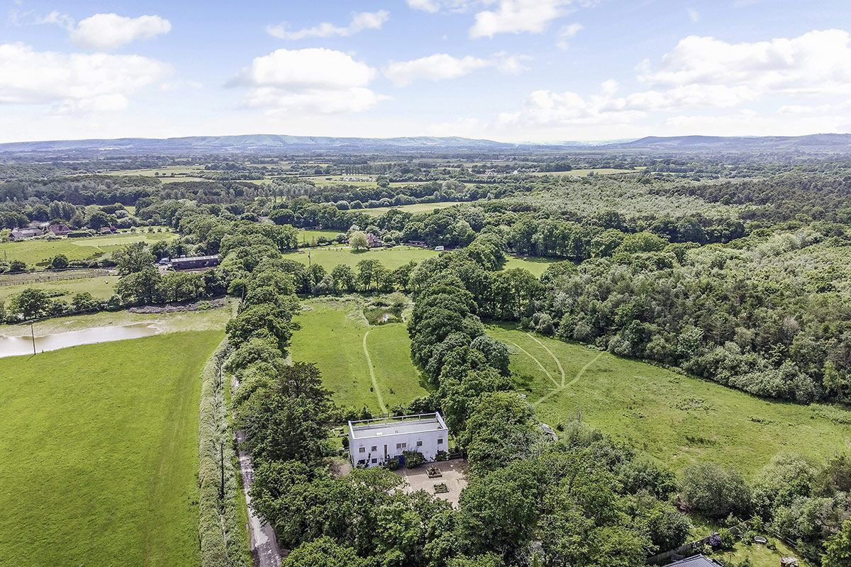 Aerial view of a white mansion surrounded by green fields and trees under a cloudy blue sky.