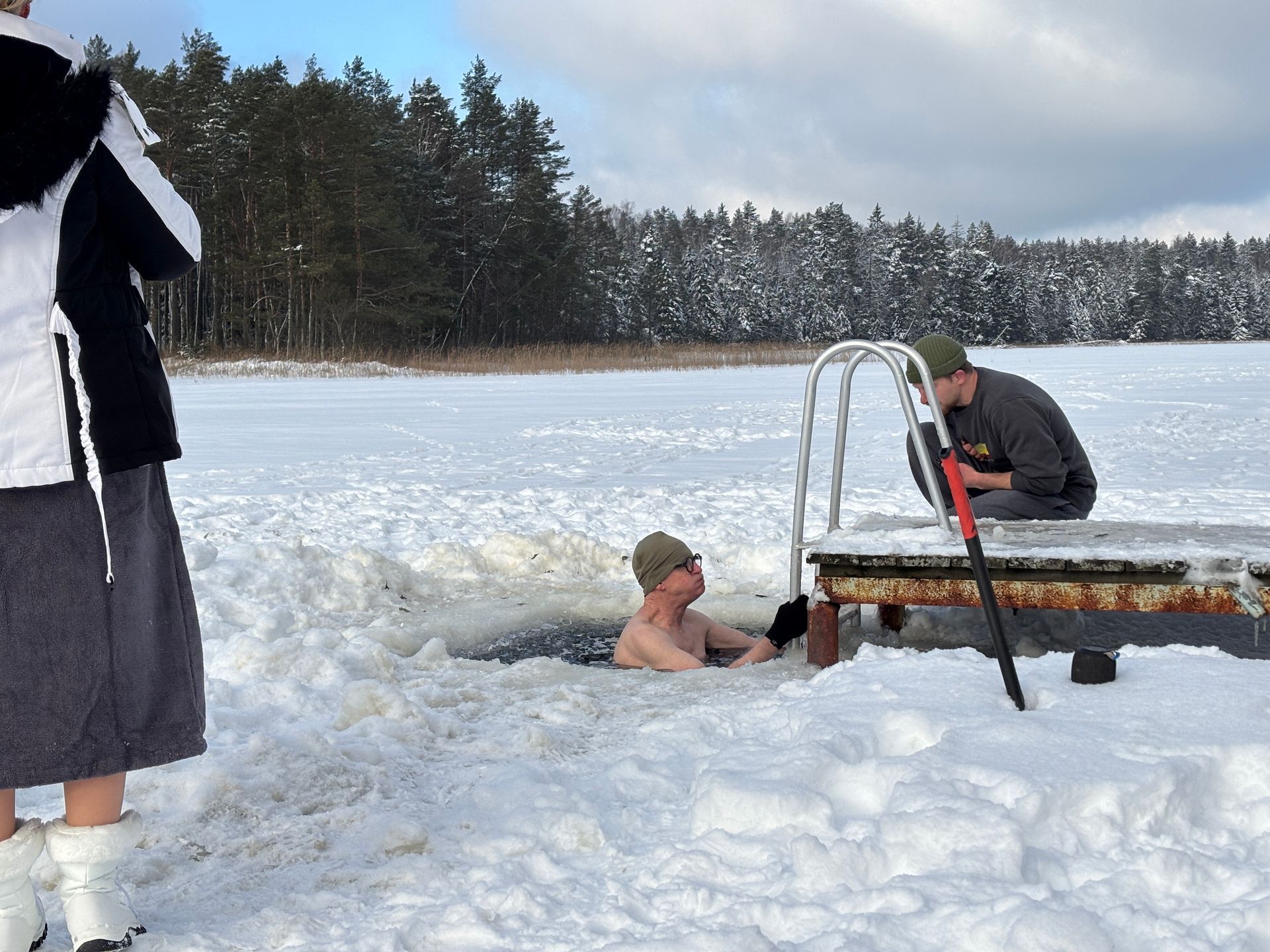 Person swimming in an ice hole, near a pier. Another person nearby. Snowy landscape, sunny day.