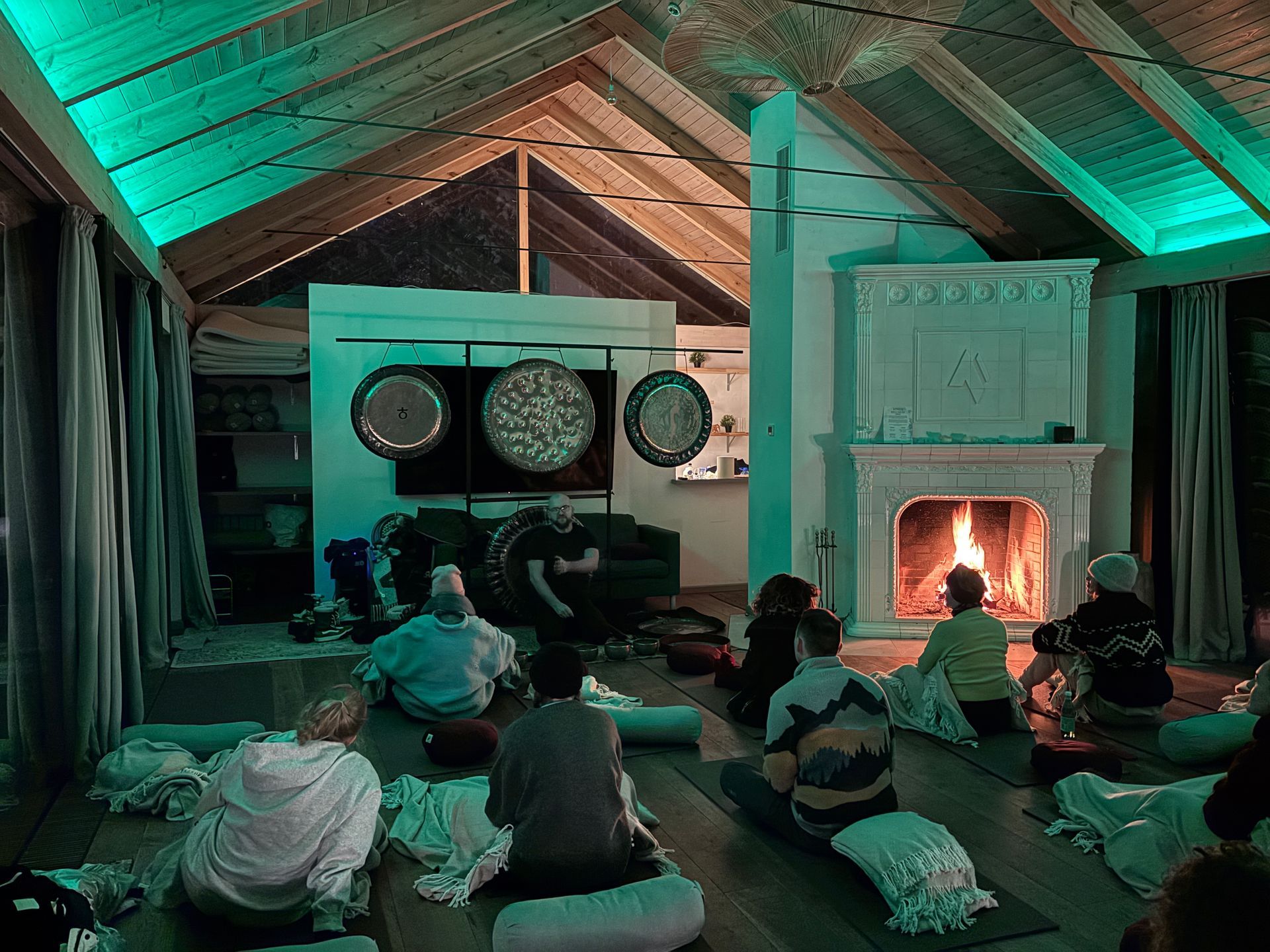 People in a yoga session, gathered near gongs and a fireplace, under green ambient lighting.