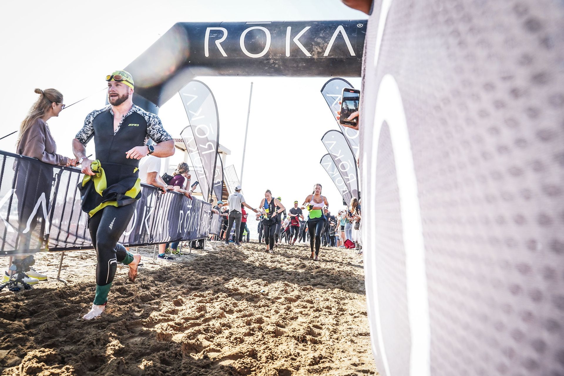 Woman with curly blonde hair in a white off-the-shoulder top, posing outdoors near green plants.