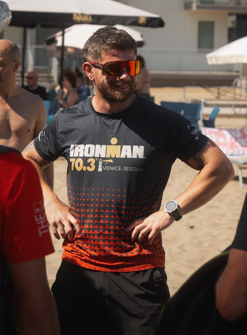 A man wearing sunglasses and an ironman shirt is standing on a beach.