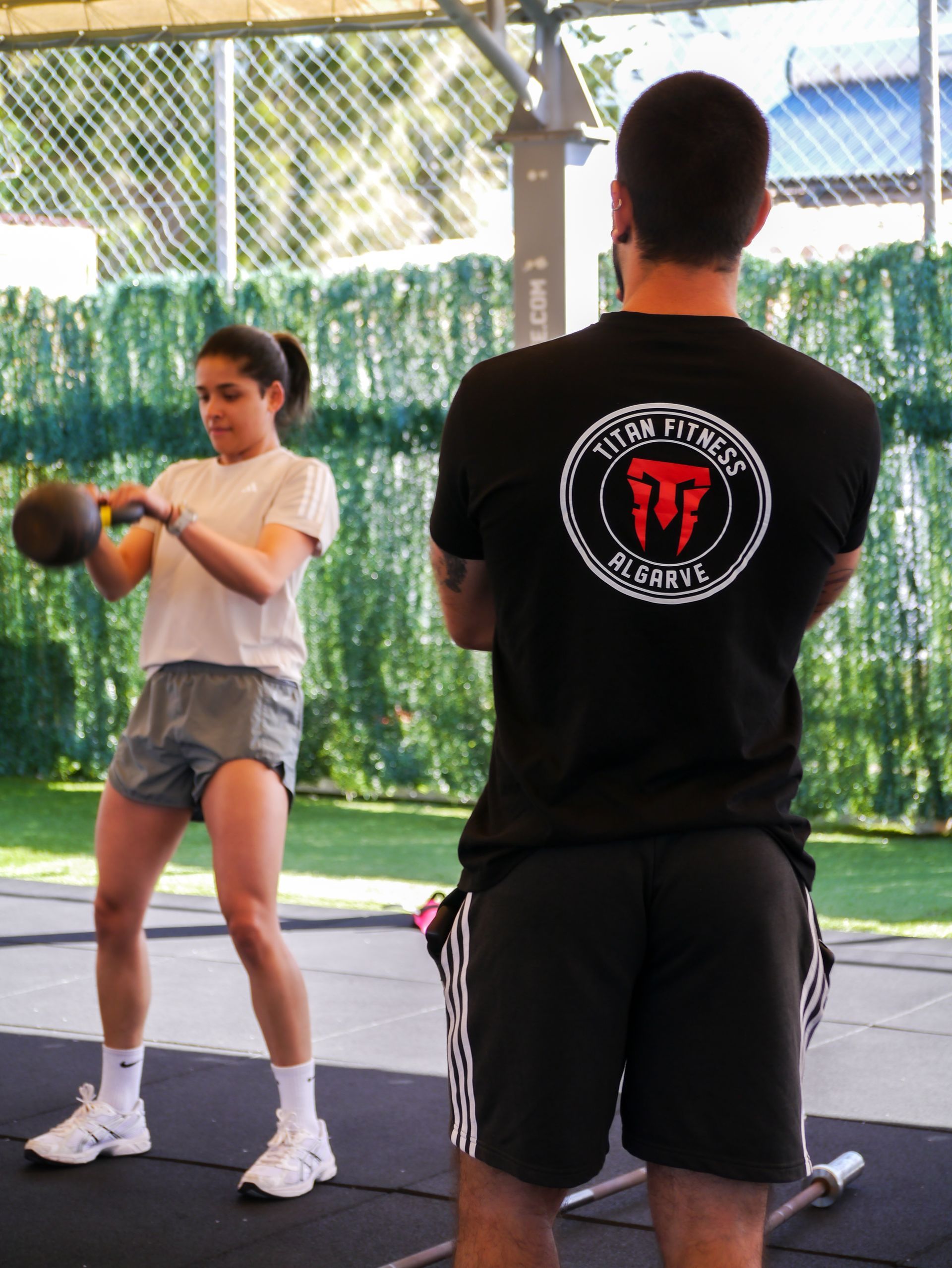 A trainer in a black branded t-shirt watches a person perform a kettlebell swing in an outdoor fitness area.