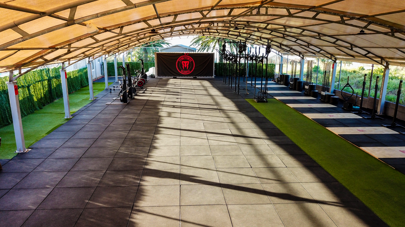 An open-air gym under a fabric canopy, featuring black rubber flooring, artificial grass strips, and exercise equipment.