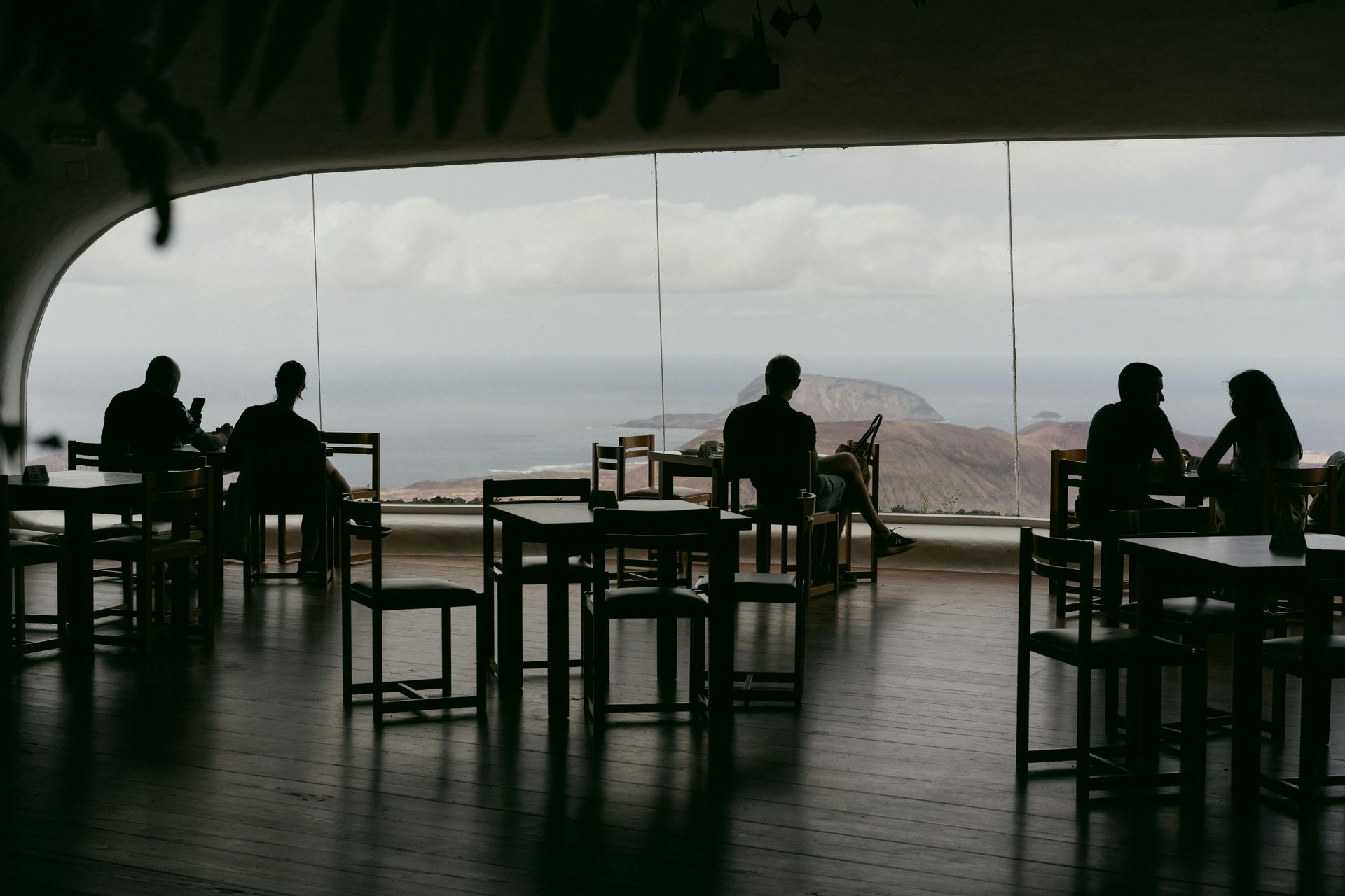 A group of people are sitting at tables in front of a window overlooking the ocean.