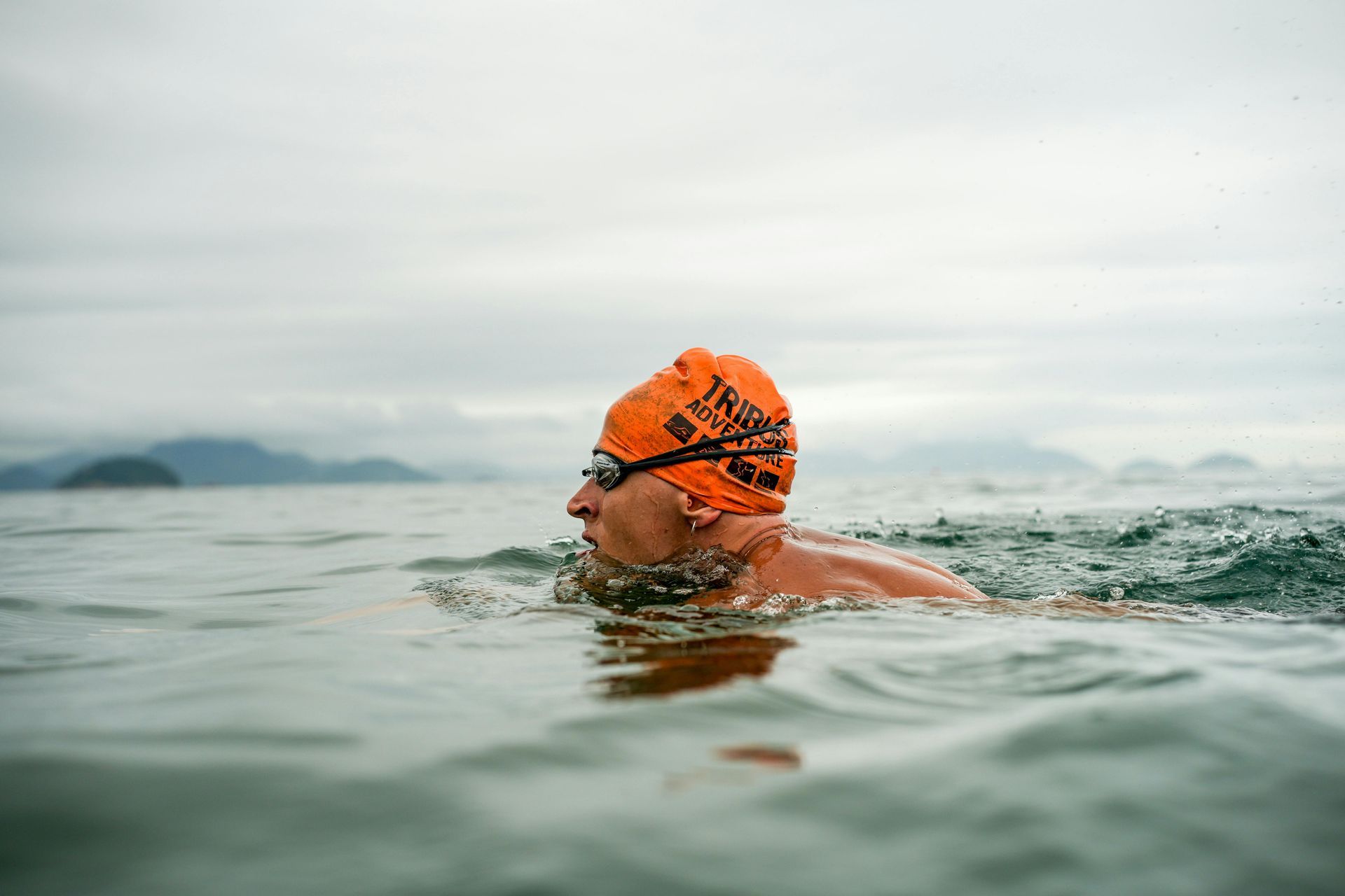 A man wearing an orange swim cap and goggles is swimming in the ocean.