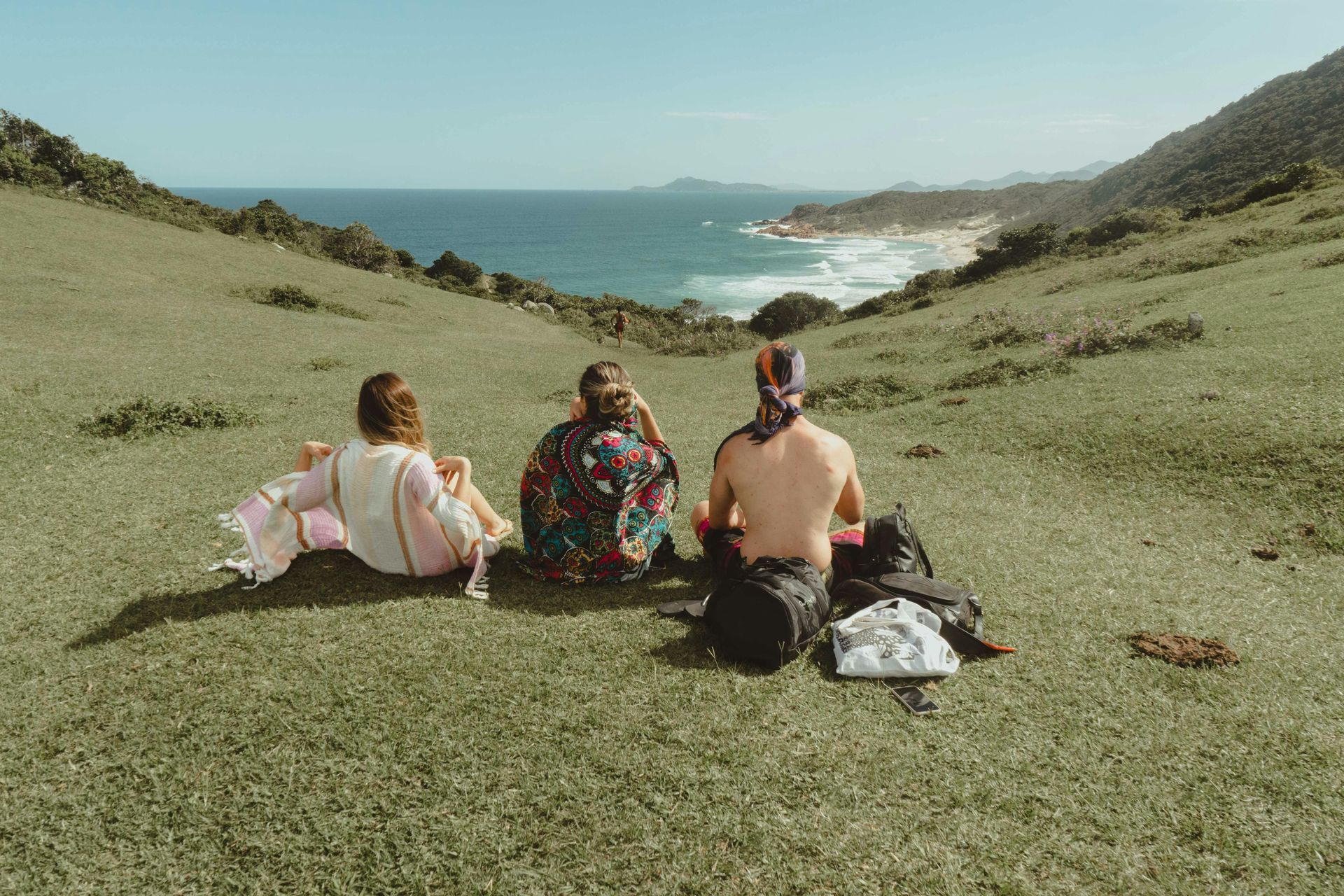 Three people are sitting on top of a grassy hill overlooking the ocean.